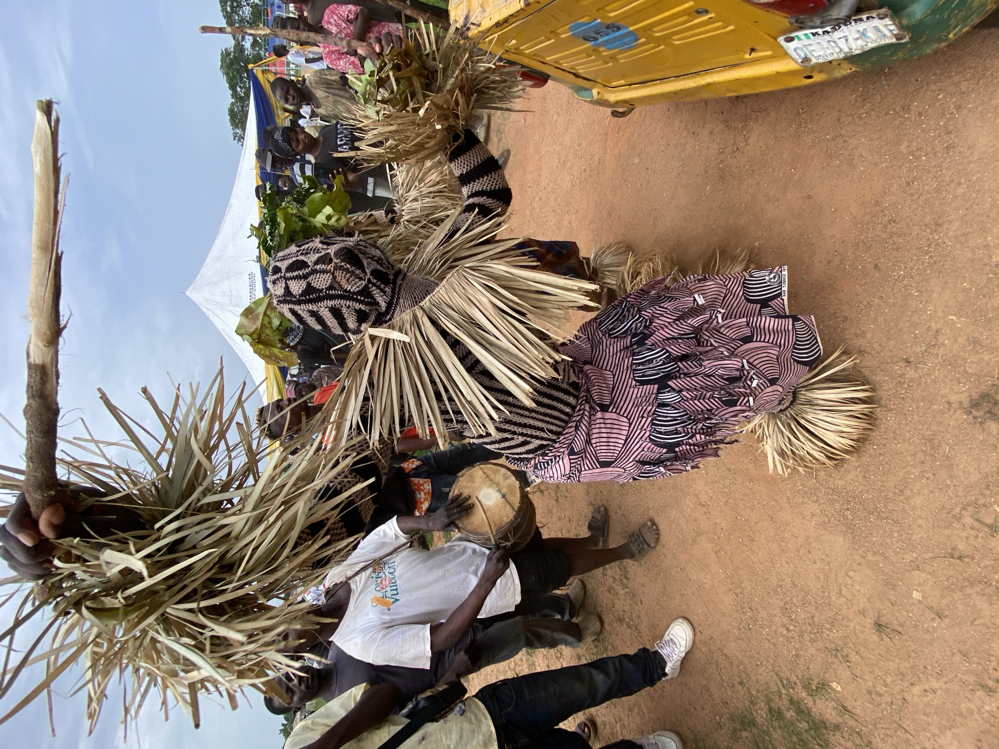 The image shows a gathering of people in what appears to be a cultural celebration in Kafanchan, Nigeria. The location is evidenced by a yellow transport vehicle bearing a license plate which reads "OE2197-KA", which indicates Kaduna State of Nigeria. Several individuals are dressed in traditional masquerade costumes made of woven straw and patterned fabrics. Some people are holding and playing traditional drums, while others are observing. A white tent is visible in the background, suggesting an organized event. The ground is dirt-covered, indicating an outdoor setting.