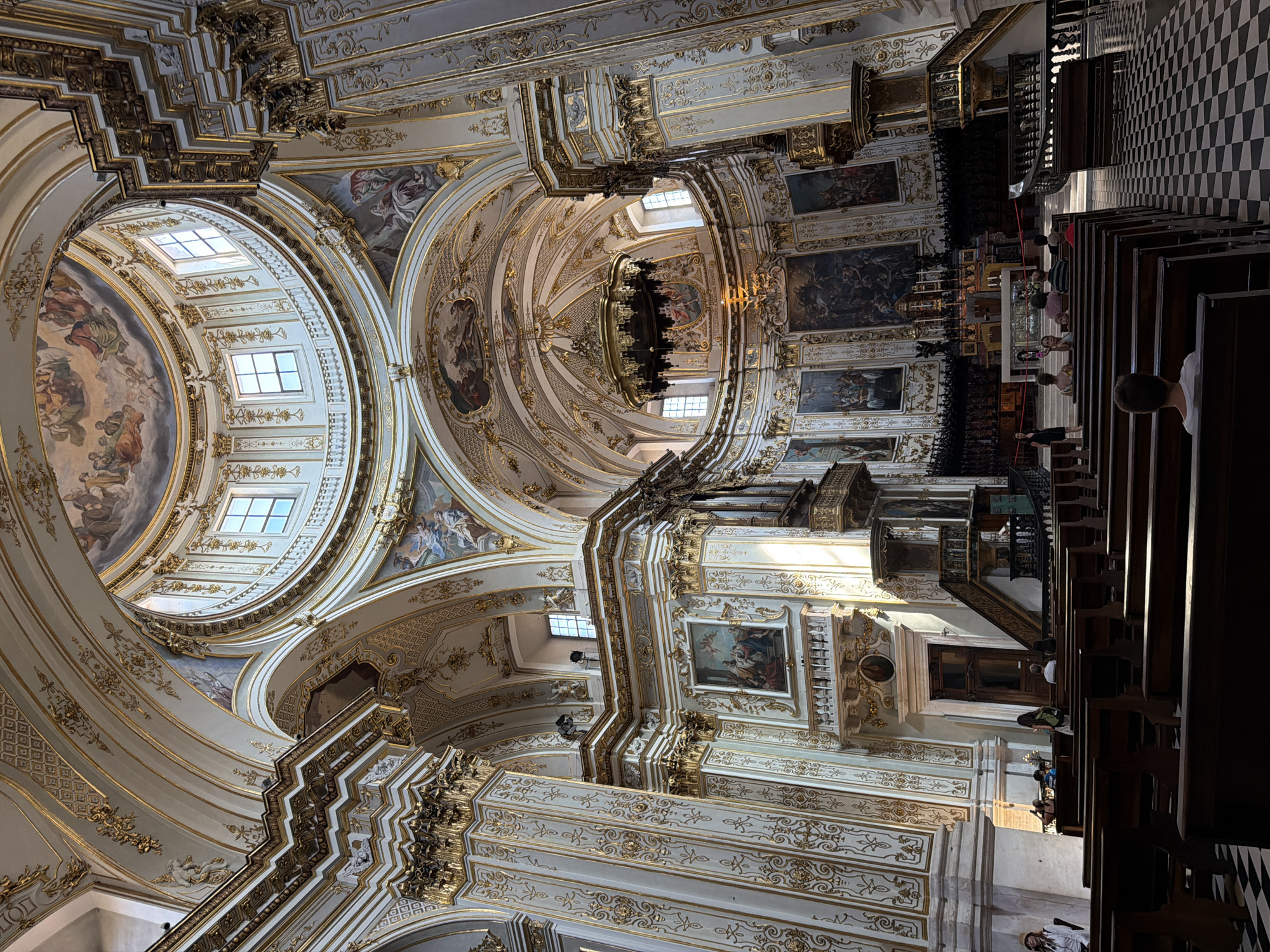 The image depicts the interior of a grand church, likely a cathedral, featuring opulent Baroque or Rococo architecture. The dominant colors are white and gold, with intricate decorative patterns adorning the walls, columns, and ceilings. A large dome is visible at the top, adorned with frescoes and illuminated by natural light from windows. Several large paintings hang on the walls, depicting religious scenes.
The foreground shows rows of dark wooden pews, some occupied by individuals. The floor is a checkered pattern of black and white tiles. In the mid-ground, there are pulpits and architectural elements typical of a church, including an altar area in the distance, though it is partially obscured.
The overall impression is one of grandeur, artistry, and religious solemnity.
Regarding the location in Mozzo, Italy: While the image is visually consistent with many Italian churches, a definitive identification of Mozzo as the specific location cannot be made solely from the visual information provided.