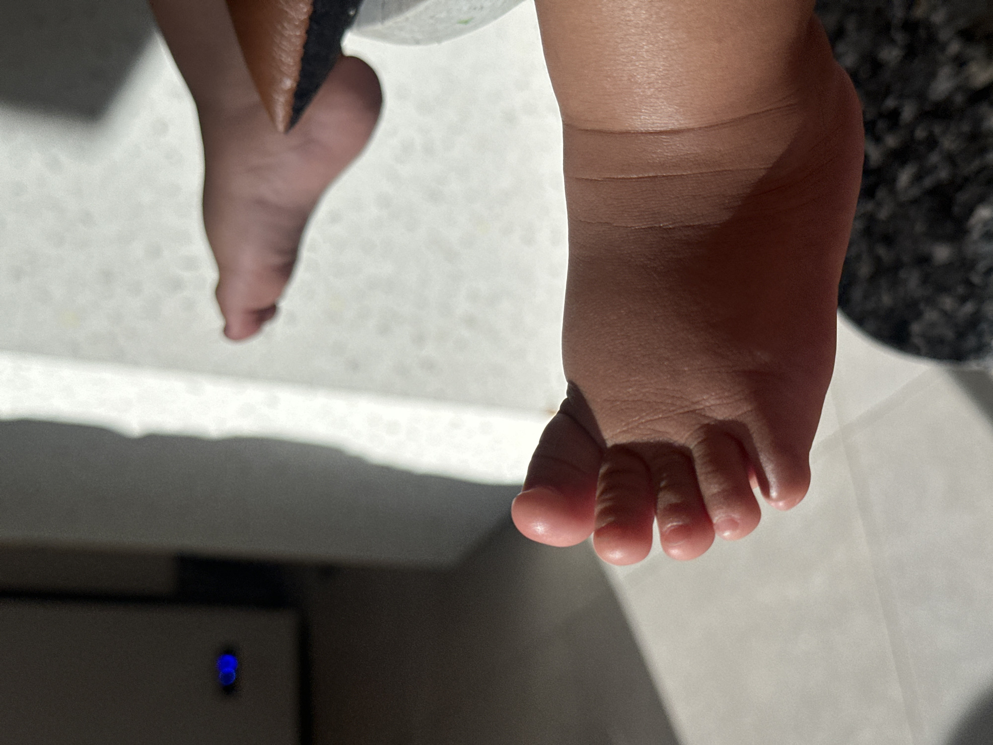The image shows a close-up view of two bare feet, likely belonging to a baby or young child. The feet are dark-skinned. The toes of the right foot are clearly visible, with small, rounded toenails. The left foot is partially obscured. The background is a light-colored surface. A portion of a dark-colored object is also visible in the image. There is also a blurry, rectangular object in the image, possibly a power outlet. The image does not have enough information to determine the location of North Kuta, Indonesia.