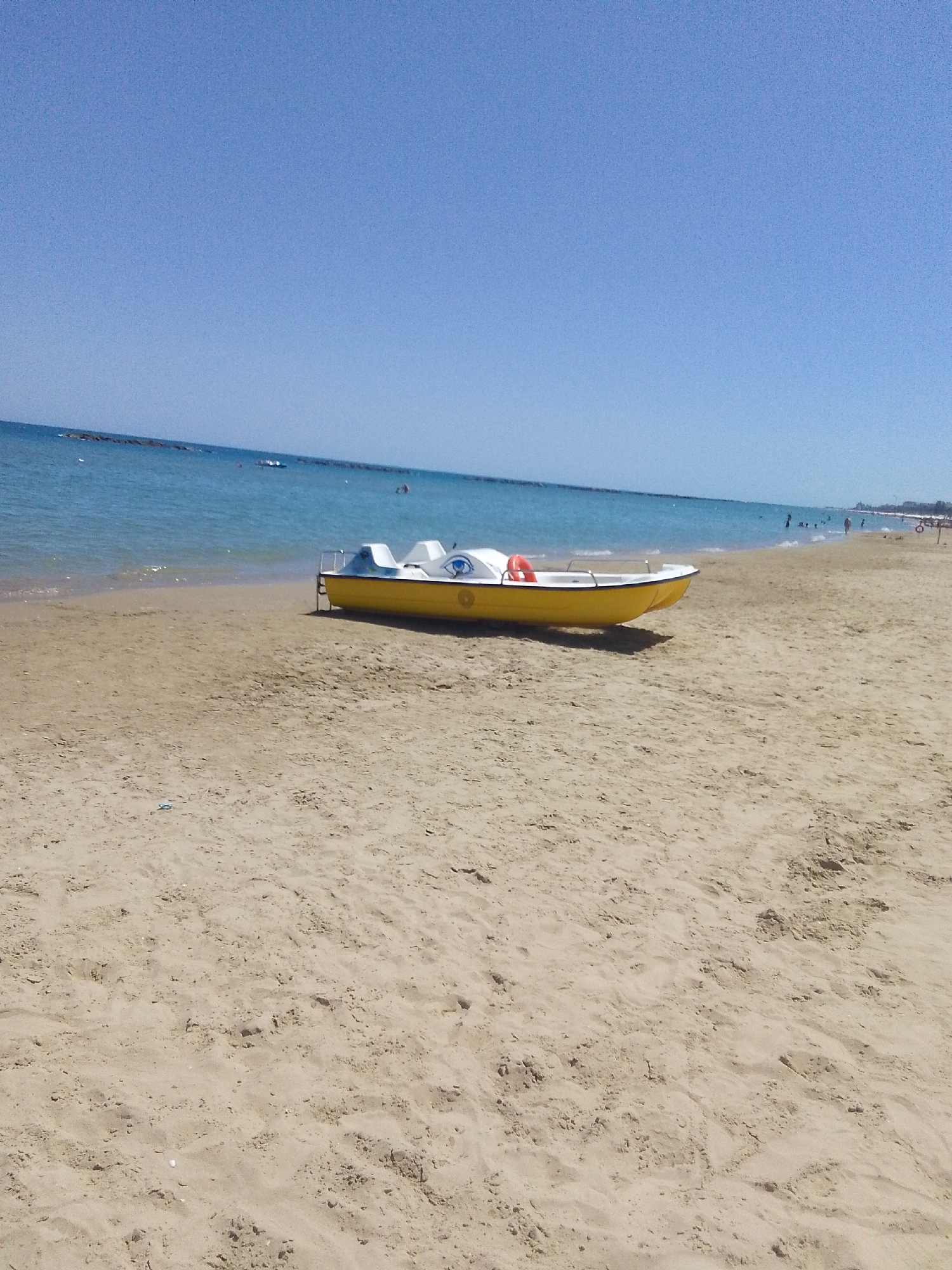 A pedal boat, yellow with white seating and a blue eye graphic, rests on a sandy beach next to the calm blue sea under a clear sky. The boat has a red life preserver attached to its side. In the distance, several people are visible in the water and on the beach, and a breakwater extends into the sea. The scene depicts a sunny day at a beach resort, likely in a coastal area of Italy.