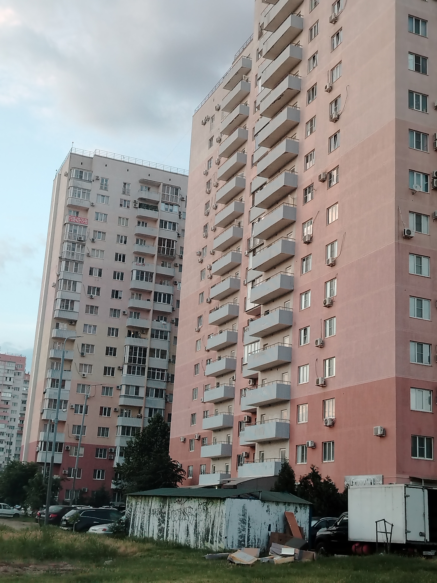 This is a medium-angle, full-frame shot of a residential area in Krasnodar, Russia. In the foreground, a patch of grass is visible, with a few pieces of debris and a partially constructed structure made of corrugated metal with graffiti. Beyond this, a line of parked cars is visible, mostly sedans and SUVs, in various shades of white, silver, black, and dark blue.
Dominating the midground and background are several multi-story apartment buildings. The building on the right is tall and painted a pale pink with darker pink accents at the base. It features numerous balconies with light gray railings and air conditioning units mounted on the exterior walls. The building on the left is slightly shorter and appears to be painted a light beige with sections of pale pink and dark red. It also has many balconies, some enclosed with windows, and air conditioning units. Another apartment building can be seen in the far background to the left, appearing even taller and lighter in color.
The sky above is overcast with a mix of light gray and pale blue clouds. The overall impression is of a typical urban residential environment, possibly during the daytime, with a slightly hazy or cloudy atmosphere. There are no people visible in the frame.
