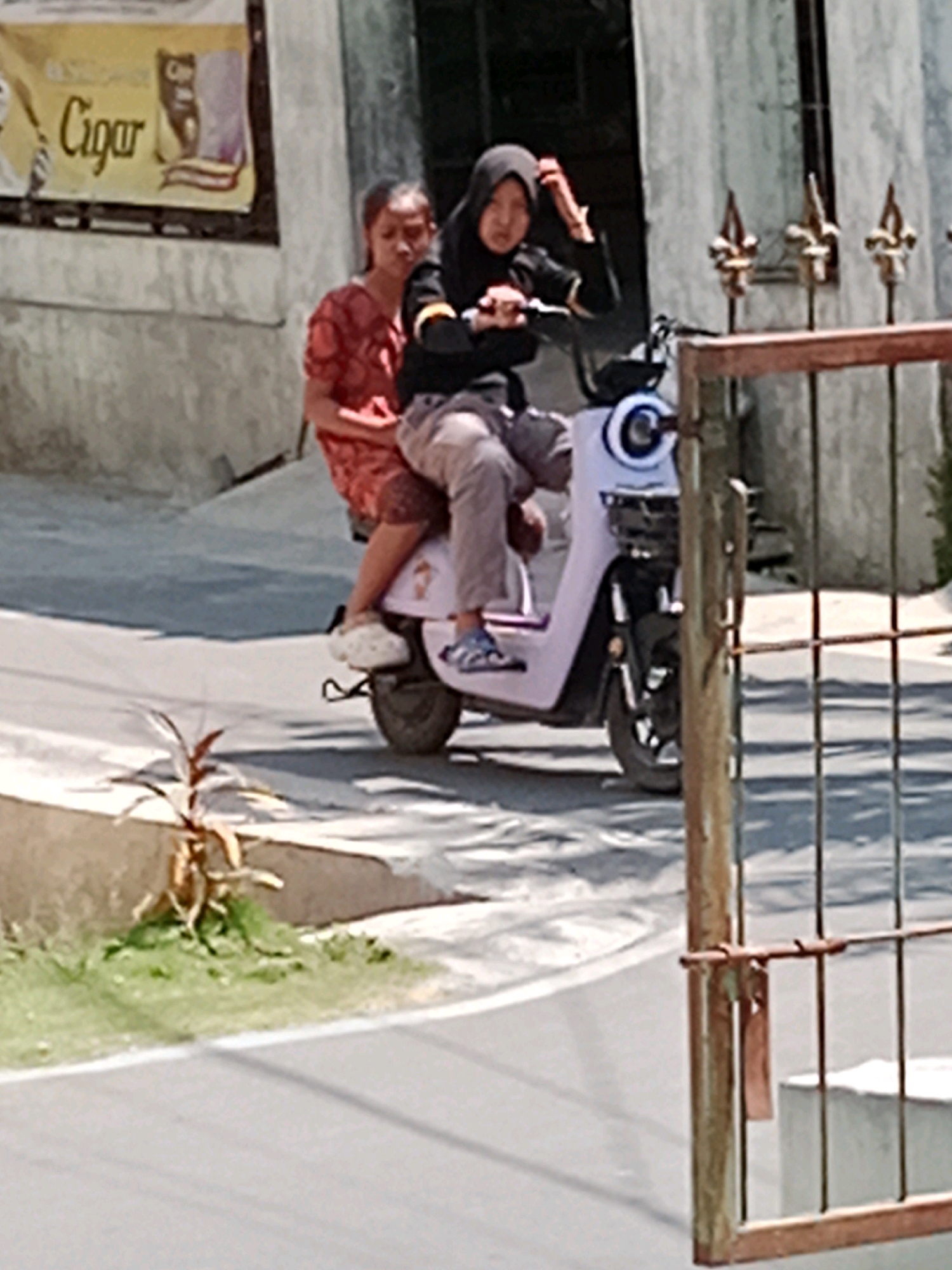 Two young females are riding a light pink electric scooter on a paved road in Waru, Indonesia. The female in the front is wearing a black hijab and a black long-sleeved shirt, and she is operating the scooter. The female in the back is wearing a red patterned top. They are both wearing casual attire, with the driver wearing gray pants and the passenger wearing light-colored pants. The scooter has a round white headlight with a black center and a basket in front. In the background, a concrete wall with a weathered banner that reads "Cigar" is visible. A rusted metal fence with vertical bars is in the foreground, partially obscuring the scooter and the road. The environment appears to be outdoors in a street setting, with some greenery and shadows present. The lighting suggests it is daytime.