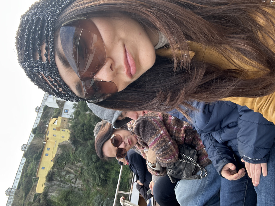 The image depicts a group of people seated on a vessel, likely on the Douro River, with the landscape of Vila Nova de Gaia, Portugal, in the background.
In the right foreground, a woman with long, dark brown hair and a dark, textured head covering wears large, brown wraparound sunglasses and has pink lipstick. She is dressed in a light-colored turtleneck and a mustard-yellow jacket. To her left, another woman is seated, wearing large, brown-rimmed round sunglasses, a multi-textured brown and pink patterned jacket, and blue jeans. Her arms are crossed, and a dark quilted handbag with a silver chain strap rests on her lap. Partially visible between these two women is a person in a grey hooded sweatshirt. Further to the left and lower in the frame, a person, likely a child, is seated, wearing a navy blue puffer jacket with "OMNI-HEAT" visible on the sleeve, and blue jeans. Their hands are resting on their lap.
The background shows a steep hillside covered with green vegetation, rising from what appears to be the riverbank. Several buildings are integrated into the slope, including a prominent yellow structure with multiple windows. Above these, a long, white, multi-storied building with a red-tiled roof is visible. Along the uppermost ridge, a distinctive long, white walled structure with evenly spaced turrets or decorative elements is visible, characteristic of the Mosteiro da Serra do Pilar in Vila Nova de Gaia. The sky is overcast.