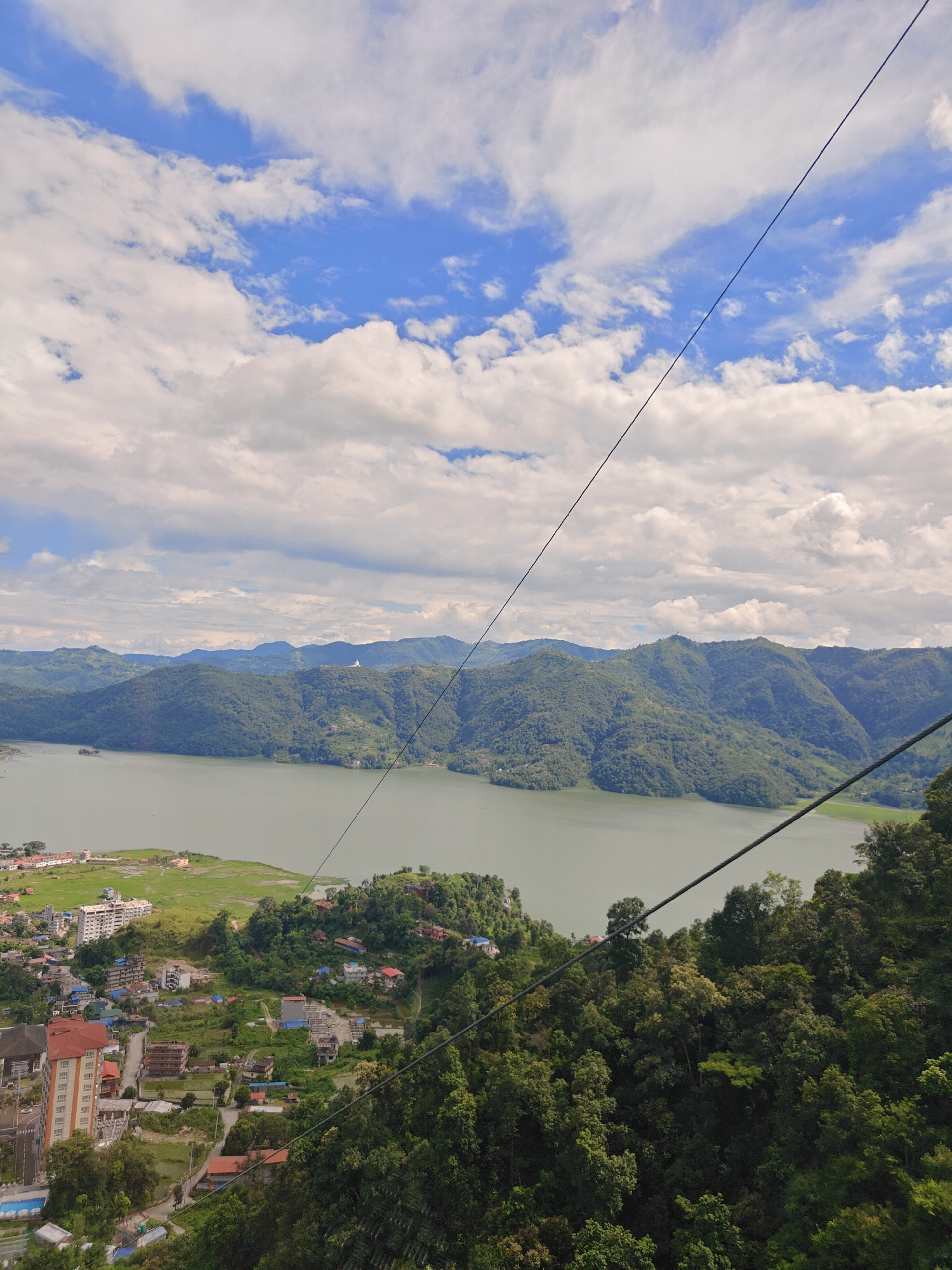 The image shows a wide-angle view of Pokhara, Nepal, dominated by the serene Phewa Lake and the lush green hills of the Annapurna range. The foreground is occupied by a dense forest with a cable car wire stretching across the frame. Below the forest, a town is visible with a cluster of buildings, including some multi-story structures and smaller houses, nestled amongst the greenery. Further into the distance, Phewa Lake stretches out, its calm waters reflecting the cloudy sky. Beyond the lake, the rolling hills, covered in vegetation, form a majestic backdrop, with hints of more distant, mist-shrouded peaks. The sky is a brilliant blue, dotted with fluffy white clouds that cast shadows on the landscape. The scene appears to be captured from a high vantage point, possibly from a cable car or a hilltop overlooking the city and the lake. The overall atmosphere is one of natural beauty and tranquility.