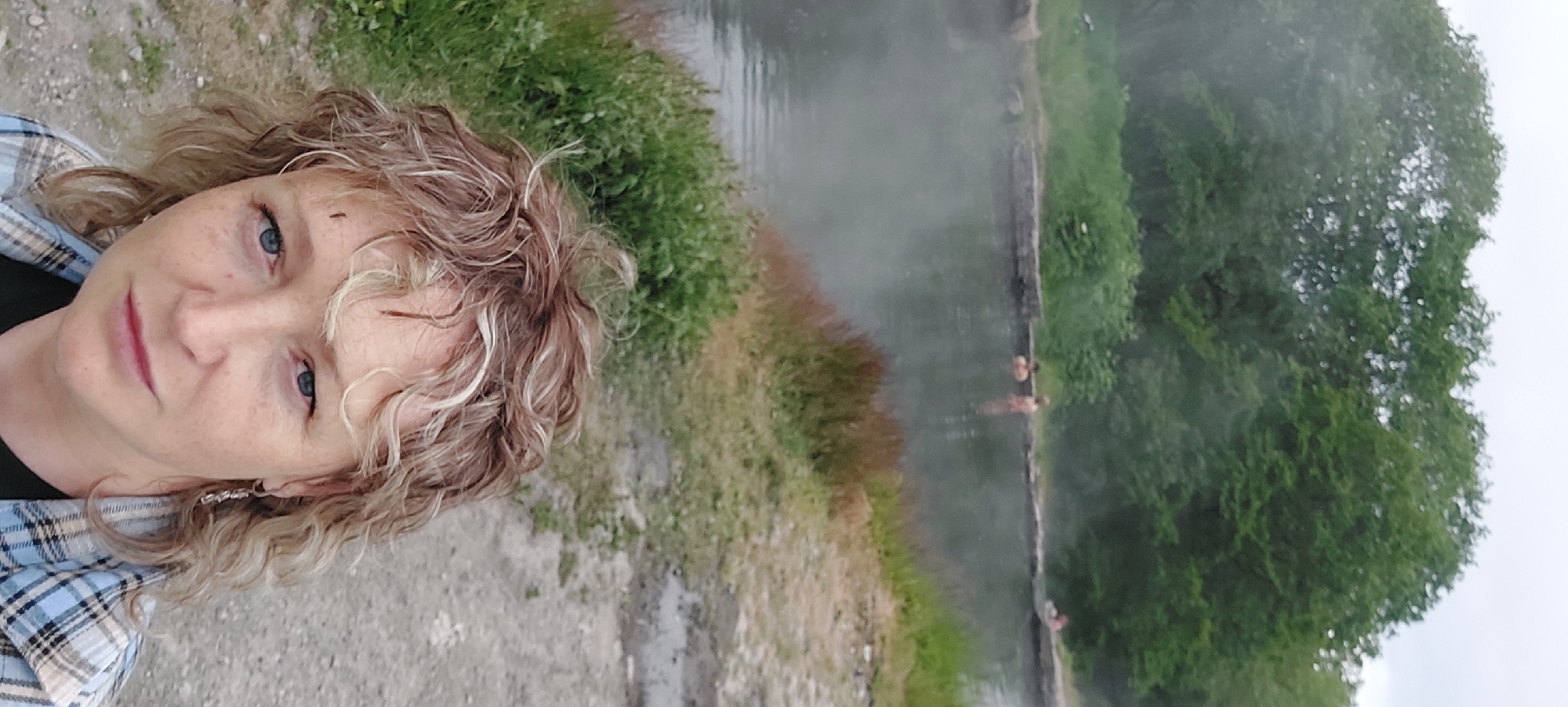 This is a selfie taken by a woman with curly blonde hair in the foreground. She is looking directly at the camera with blue eyes and has freckles on her face. She is wearing a plaid shirt over a dark top and an earring in her left ear. In the background, there is a body of water with steam rising from it, suggesting it may be a hot spring. Two people are visible in the water in the middle ground, and another person is further in the distance on the right. The water is bordered by a stone edge and surrounded by green grass and trees. The overall environment suggests an outdoor recreational area, possibly a natural hot spring located in or near Nachiki, Russia, given the presence of steam and a natural landscape.