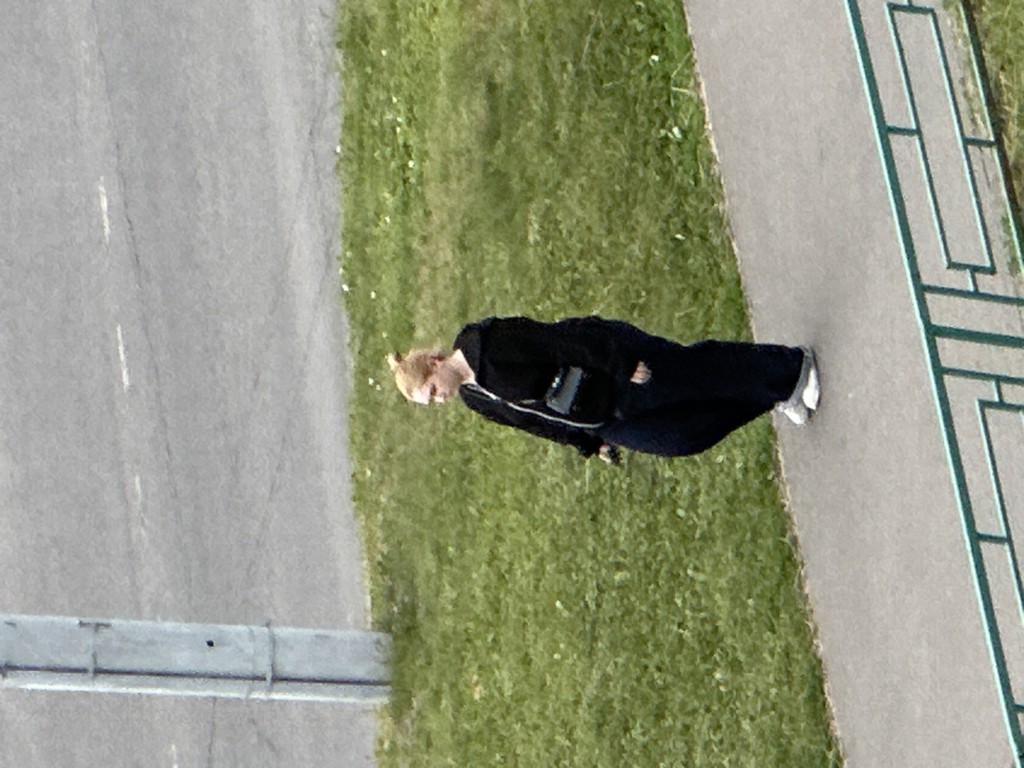 A full-body, high-angle, medium shot captures a woman walking on a sidewalk. She is wearing a black zip-up hooded jacket over dark blue pants and white sneakers. A small black crossbody bag is slung across her chest. Her blonde hair is pulled back in a bun. She is positioned on a paved sidewalk that runs alongside a grassy area. Behind the grass is a paved road with faint white markings. A concrete utility pole stands on the left side of the image, partially obscuring the road. A light green metal fence runs along the bottom of the frame, separating the sidewalk from another area. The scene is outdoors, likely in the city of Podolsk, Russia, on a day with ambient light but no direct sun.