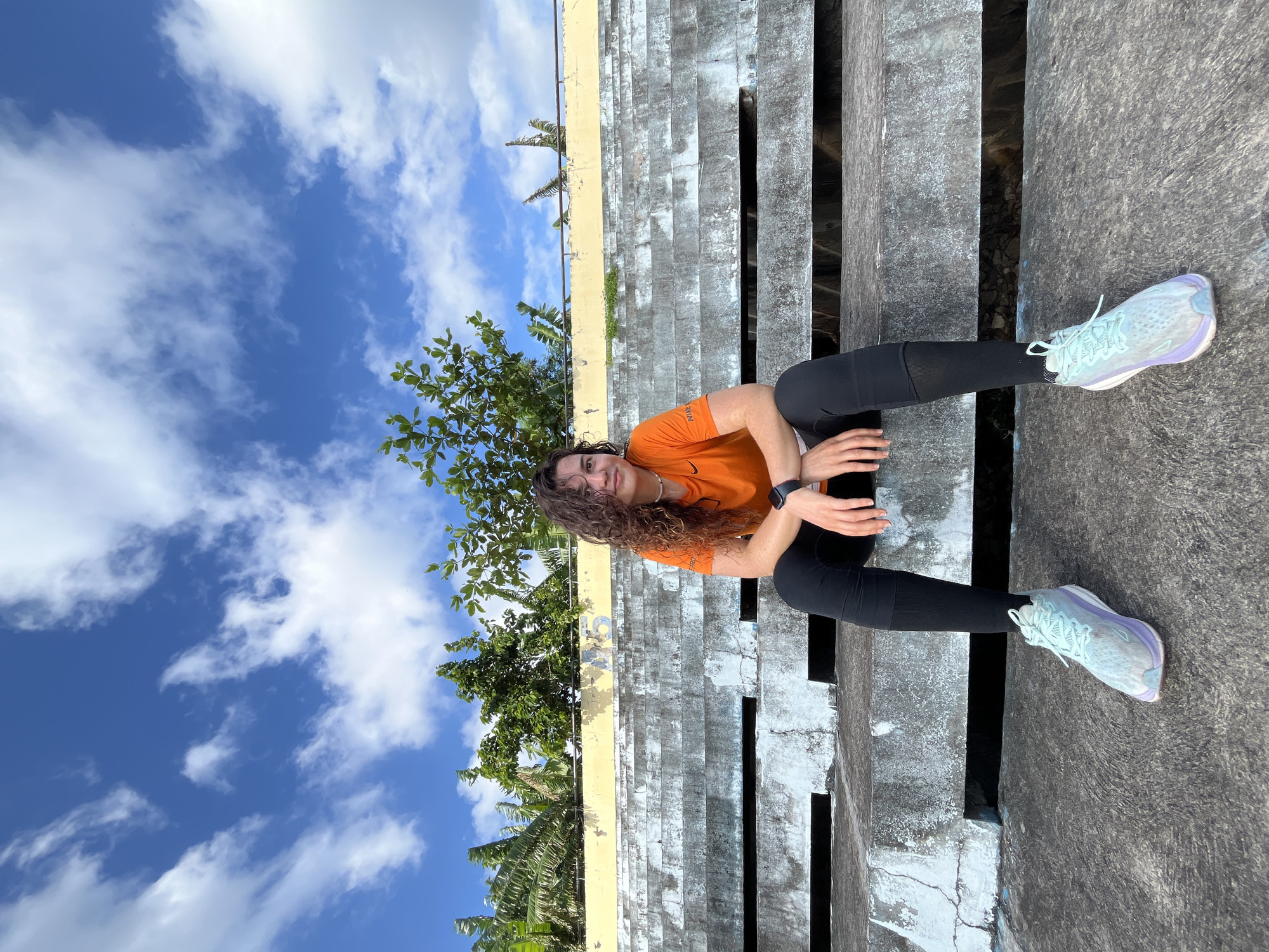 A young woman with curly brown hair, wearing an orange t-shirt, black leggings, and light blue athletic shoes, is sitting on concrete bleachers outdoors. She is looking at the camera with a slight smile. The bleachers are weathered and have some visible cracks and discoloration. Behind her, there is a yellow wall with some faded markings, and lush green trees and banana plants. The sky above is blue with scattered white clouds. The scene appears to be an outdoor sports or event venue, possibly located in Phan Thiết, Vietnam, given the tropical vegetation.