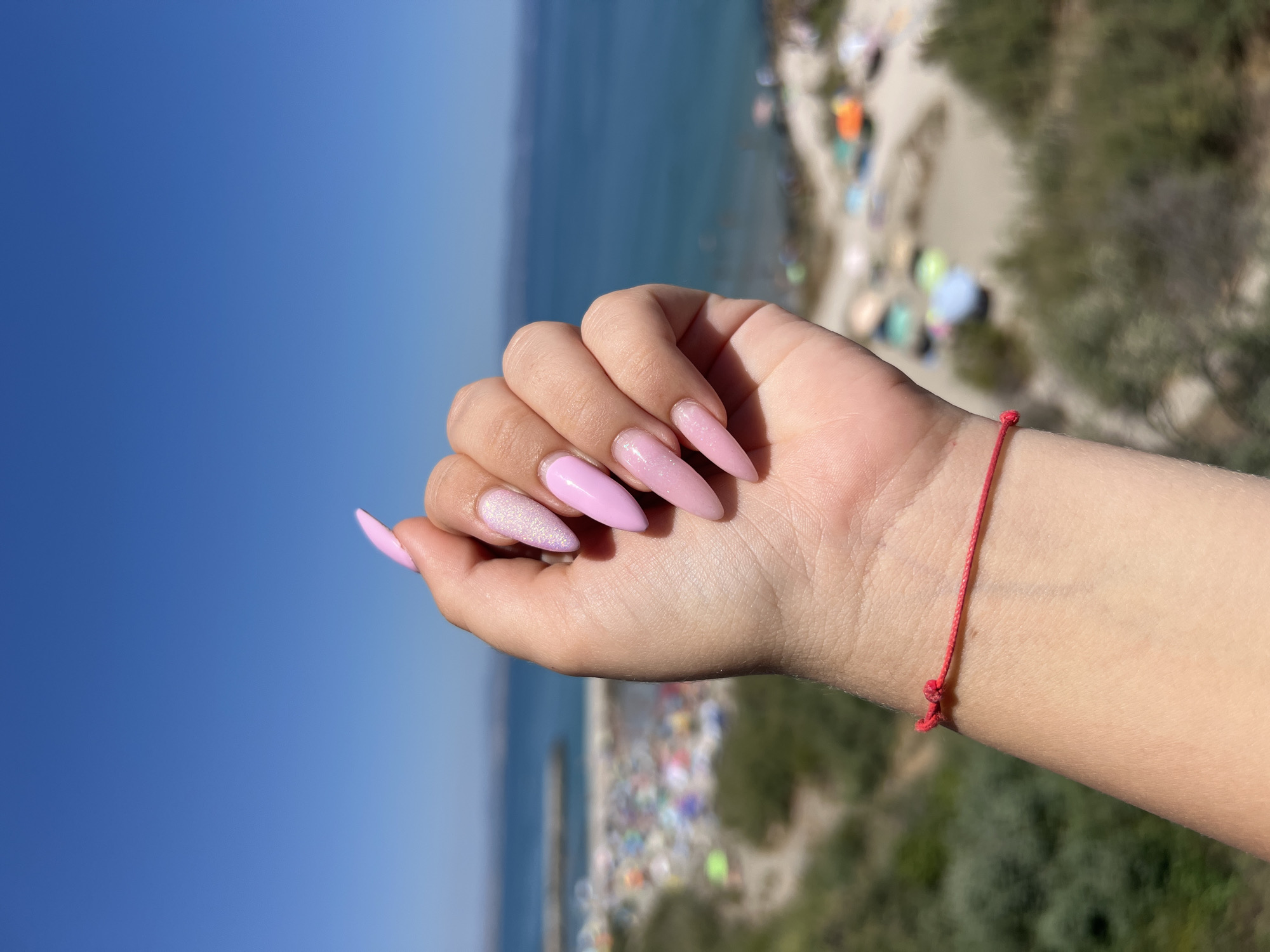 A close-up, eye-level shot features a person's left hand with long, almond-shaped nails painted light pink, some with a glittery finish, against a blurred background of the beach in Pomorie, Bulgaria. The hand is positioned in the foreground, with the palm facing the camera and the fingers slightly curled. A thin red string is tied around the wrist, just above the hand.
The background depicts a bustling beach scene under a clear blue sky. Numerous beach umbrellas and towels in various colors are scattered across the sand. The sea, a deep blue, stretches out to the horizon, meeting the sky. A breakwater or pier is visible in the distance. Green shrubs and vegetation line the edge of the beach. The overall impression is a sunny day at the seaside.