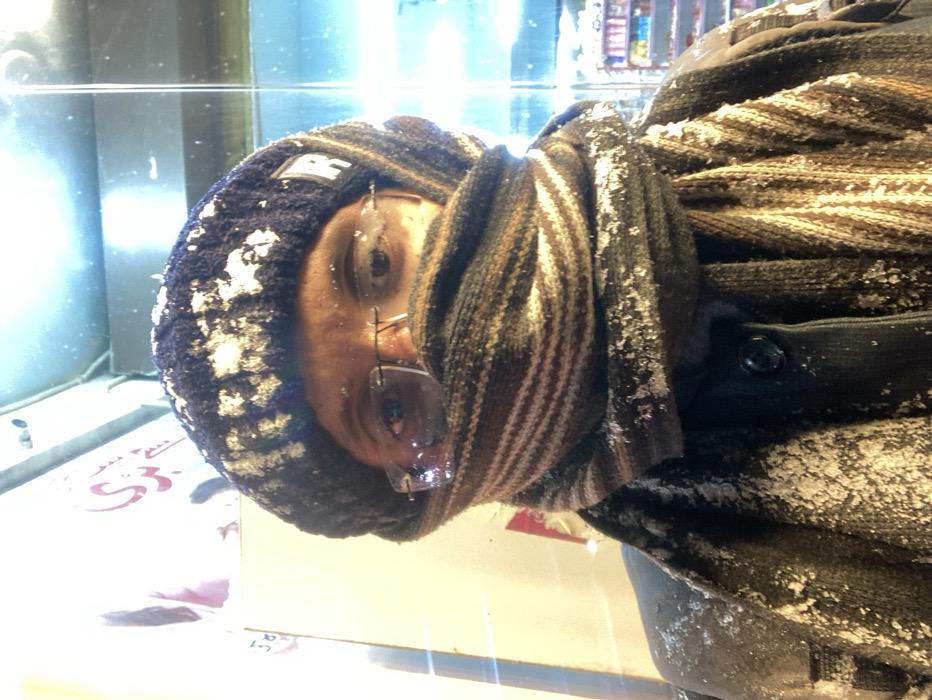 The image displays a person dressed in heavy winter clothing, oriented such that their face is seen from the side, looking slightly upwards. They are wearing a dark blue knit beanie or hat, adorned with visible white specks of snow. A small, light-colored logo or text is present on the hat. A striped scarf, featuring brown, beige, and dark brown tones, is wrapped around their neck and lower face, partially obscuring their chin and mouth. Clear-framed glasses rest on their face, with the lenses appearing somewhat fogged or iced. A dark-colored coat or jacket, also dusted with snow, is visible on their torso.
The presence of snow on the person's attire indicates cold, snowy weather, aligning with the winter climate typical of Quetta City Tehsil, Pakistan. The activity captured appears to be the person observing their surroundings or looking into a display, possibly through a window.
The background features bright, artificial lighting and reflections on a glass-like surface. Blurred elements suggest an indoor or sheltered urban environment, possibly a shop interior with shelves or products. Some indistinct text in an Arabic script, potentially Urdu or Pashto, and numbers like "53" and "51" are partially visible in the lower part of the frame. The person's expression is neutral, suggesting observation.