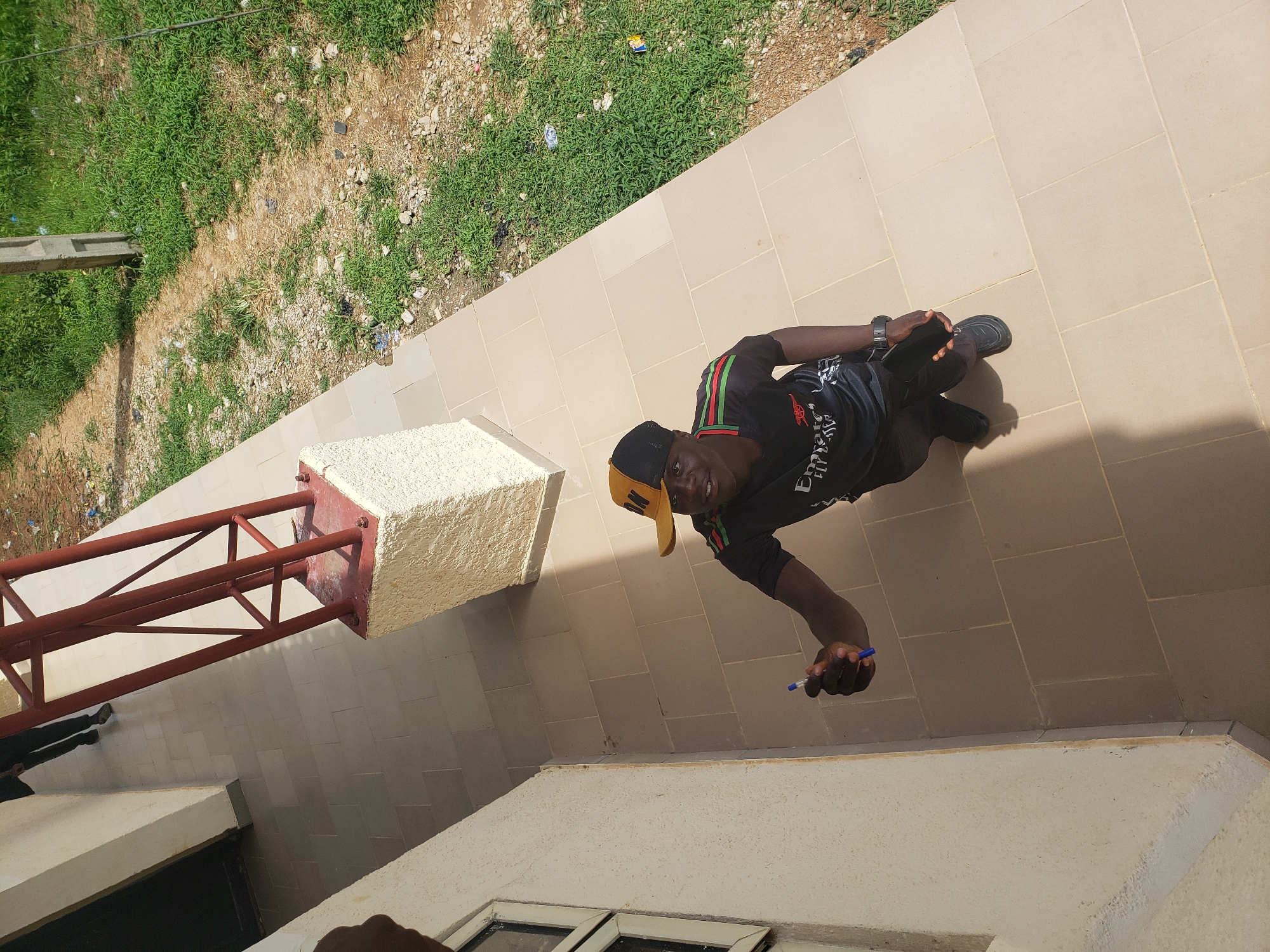 A young Black male wearing a black and yellow baseball cap, a black t-shirt with red and green stripes on the sleeves, black pants, and black shoes stands on a light brown tiled walkway next to a cream-colored building. The male is holding a blue pen in his right hand and a black smartphone in his left. He is looking up and to his right, with a slight smile. The walkway is bordered by a grassy area with some dirt patches and scattered litter. In the background, a red metal structure stands on a white concrete base, and a section of the building's wall with a window is visible. The scene appears to be in an outdoor urban environment.