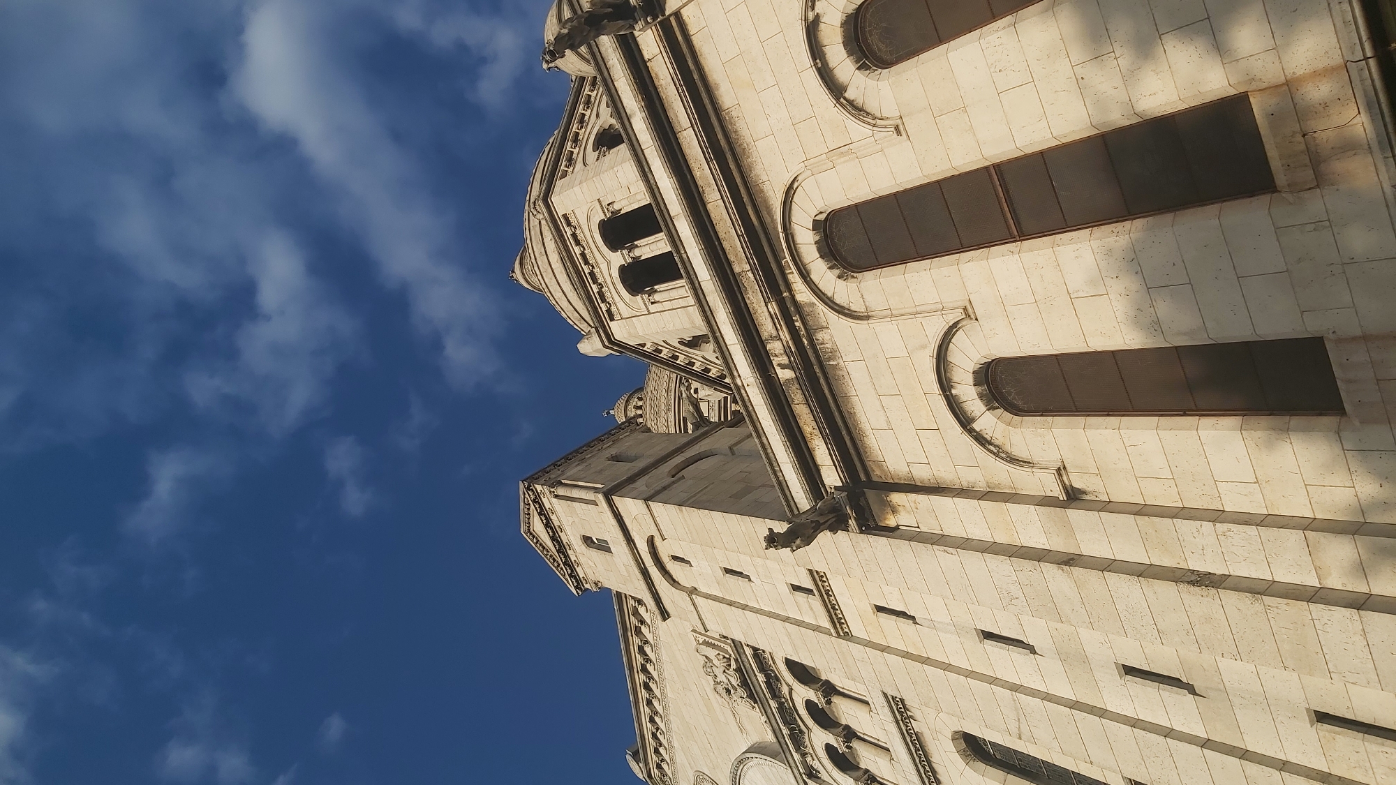 The image shows an upward view of the Sacré-Cœur Basilica in Montmartre, Paris, France. The basilica is constructed of white stone and features distinctive Roman-Byzantine architecture, including domes, arches, and intricate carvings. The sky above is a deep blue with scattered white clouds. The viewpoint emphasizes the scale and grandeur of the building, with the camera angled up towards its towers and domes. The stone walls are detailed with rectangular blocks and feature tall, arched windows. Gargoyles are visible projecting from the building. Sunlight casts shadows on the facade, highlighting the textures and architectural details. The composition is vertical, with the basilica occupying the majority of the frame against the backdrop of the sky.