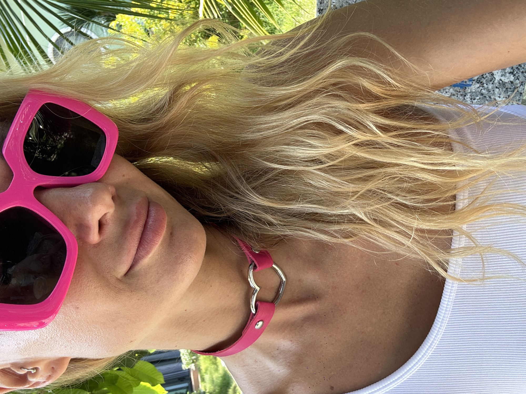 The image features a close-up, sideways shot of a woman with blonde hair and a pink choker. She is wearing large, bright pink sunglasses and a white ribbed tank top. Her skin appears tanned, and the lighting suggests it is daytime, possibly sunny, given the strong highlights on her hair and skin. She is lying down, with her head slightly tilted.
In the background, blurred green foliage and palm fronds are visible, indicating an outdoor setting with tropical vegetation. The overall mood is relaxed and summery. The woman has a subtle smile and her lips are painted with a rosy-pink lipstick. The choker is pink with a silver heart-shaped charm. The image orientation is rotated, with the woman's head to the left. There is no visible text in the image. The location context provided is Mũi Né, Vietnam, which aligns with the tropical scenery.