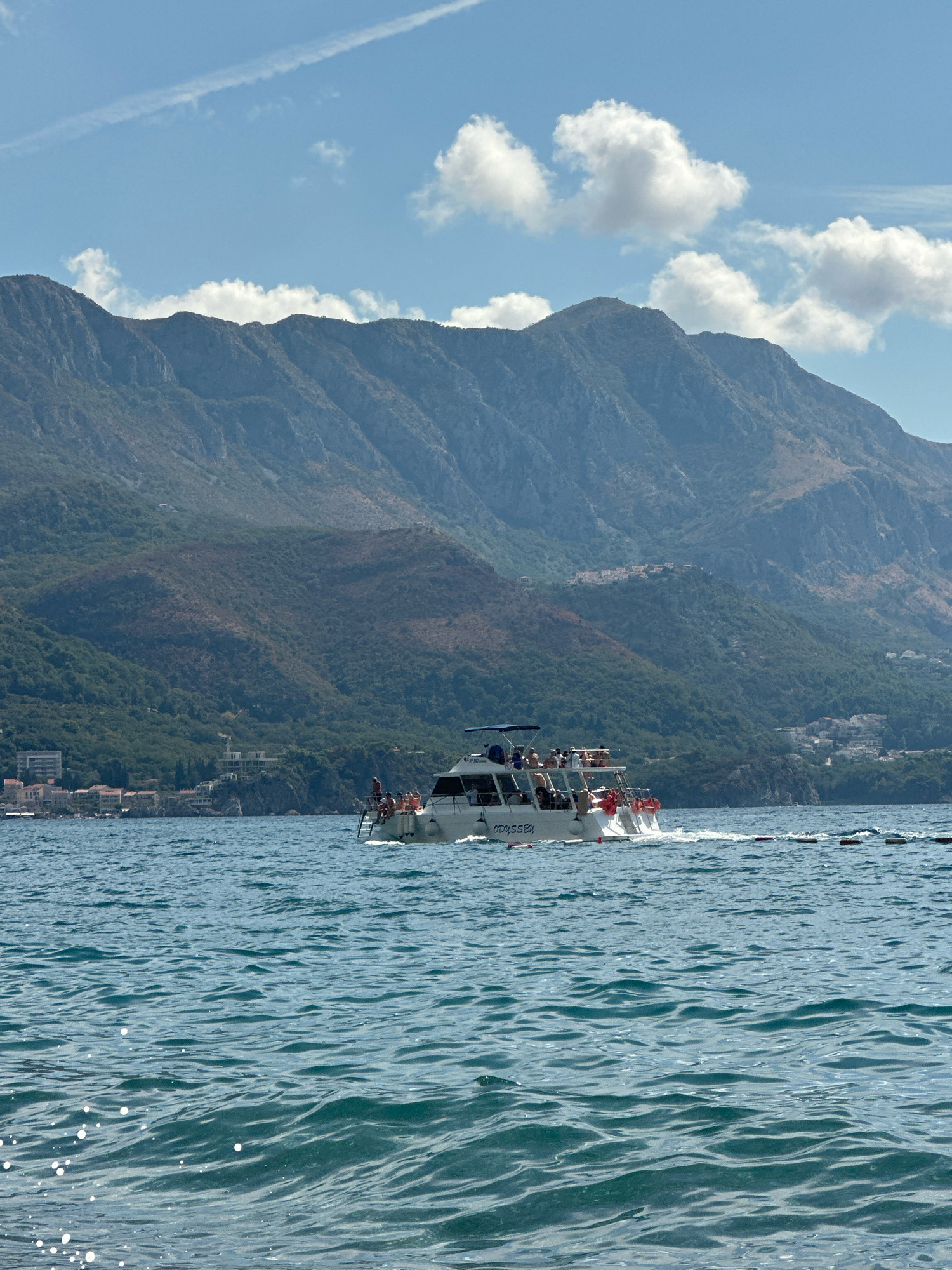 A wide shot captures a boat sailing on the Adriatic Sea with mountains in the background. The boat is white with a blue awning and appears to be carrying passengers. The sea is a deep blue with whitecaps, suggesting a slightly choppy condition. The mountains behind the sea are steep and covered in greenery, with some buildings visible on their slopes. The sky is blue with scattered white clouds, and a faint contrail is visible. The image appears to be taken from the water level, with the boat moving away from the viewer. The location appears to be near the city of Boreti, Montenegro, based on the distinctive mountainous coastline and the architecture of the buildings on the shore.
