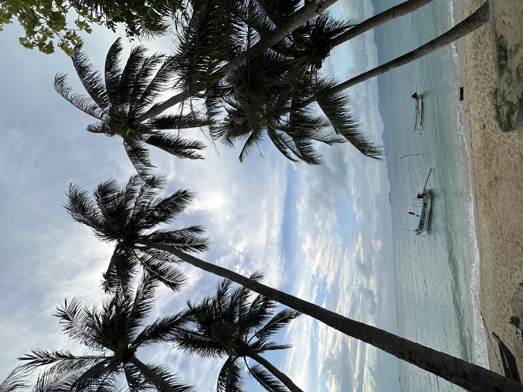 The image depicts a tranquil beach scene in the Ko Samui District of Thailand. Several palm trees frame the view, their fronds silhouetted against a cloudy sky with patches of blue. The gentle waves of the ocean lap onto a sandy shore. Two small fishing boats are visible in the water, one closer to the shore and the other further out. In the distance, the outline of mountains can be seen against the horizon. The overall atmosphere is serene and peaceful.