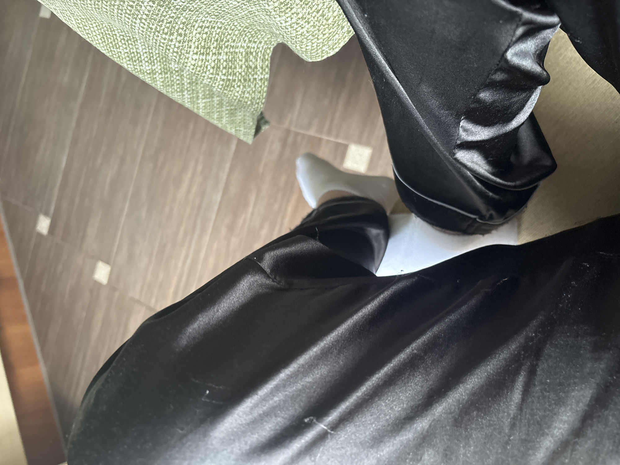 The image shows a close-up of a person's legs, wearing black satin pants and white socks. The person is sitting on a floor that appears to be tiled with a wood-grain pattern. To the right of the person's legs is a textured green fabric, possibly a blanket or upholstery. The lighting suggests an indoor environment. There is no visible information or context within the image to suggest the location is the city of Sinyavino, Russia.