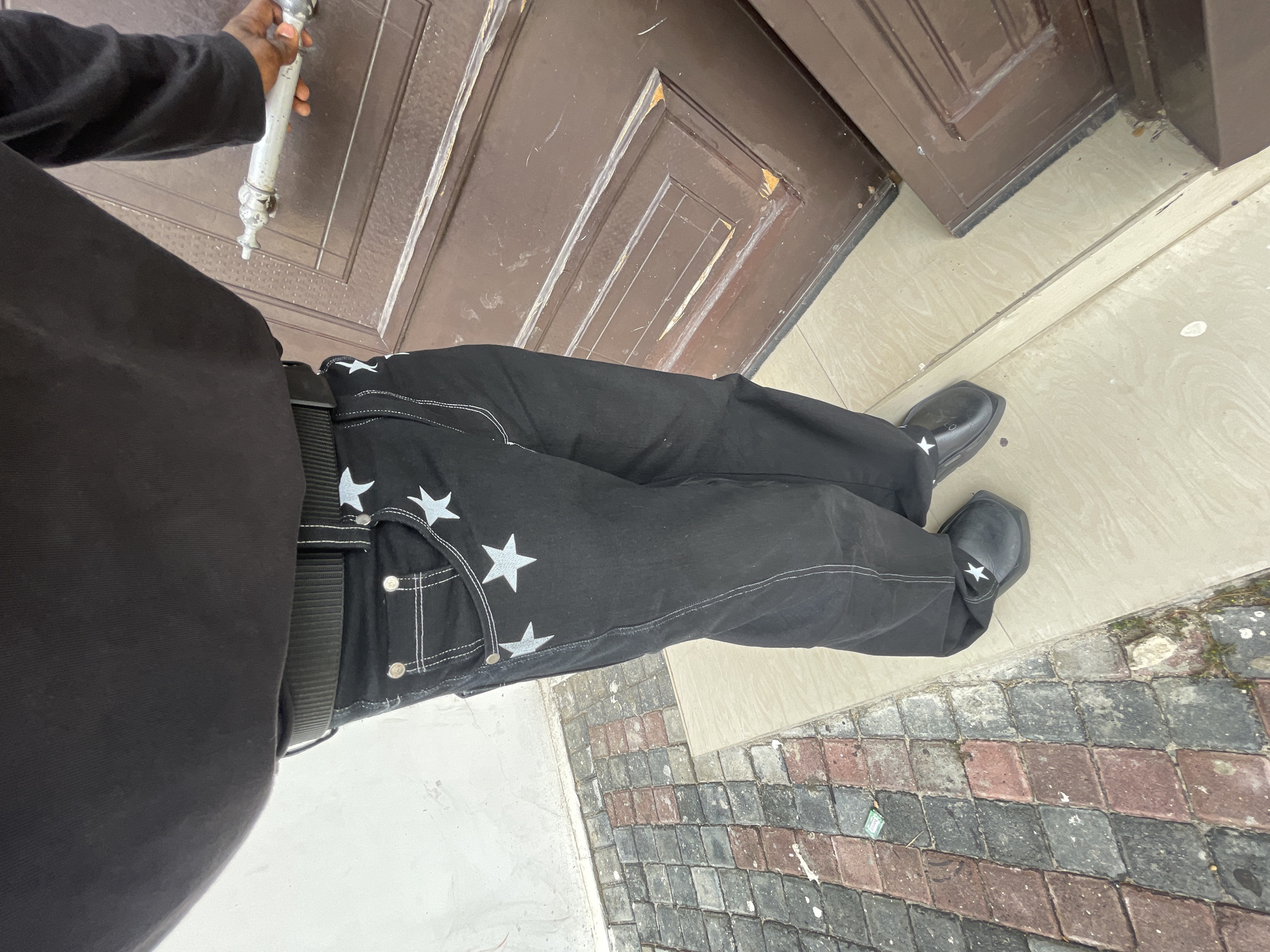 A person wearing black clothing and black pants with white star appliques is standing on a sidewalk in Lagos, Nigeria. The sidewalk is composed of irregularly shaped bricks. The person is holding a silver metal object, possibly a door handle or tool, with their right hand. A brown door is partially visible behind the person. The person is wearing black square-toed shoes, each with a white star applique.