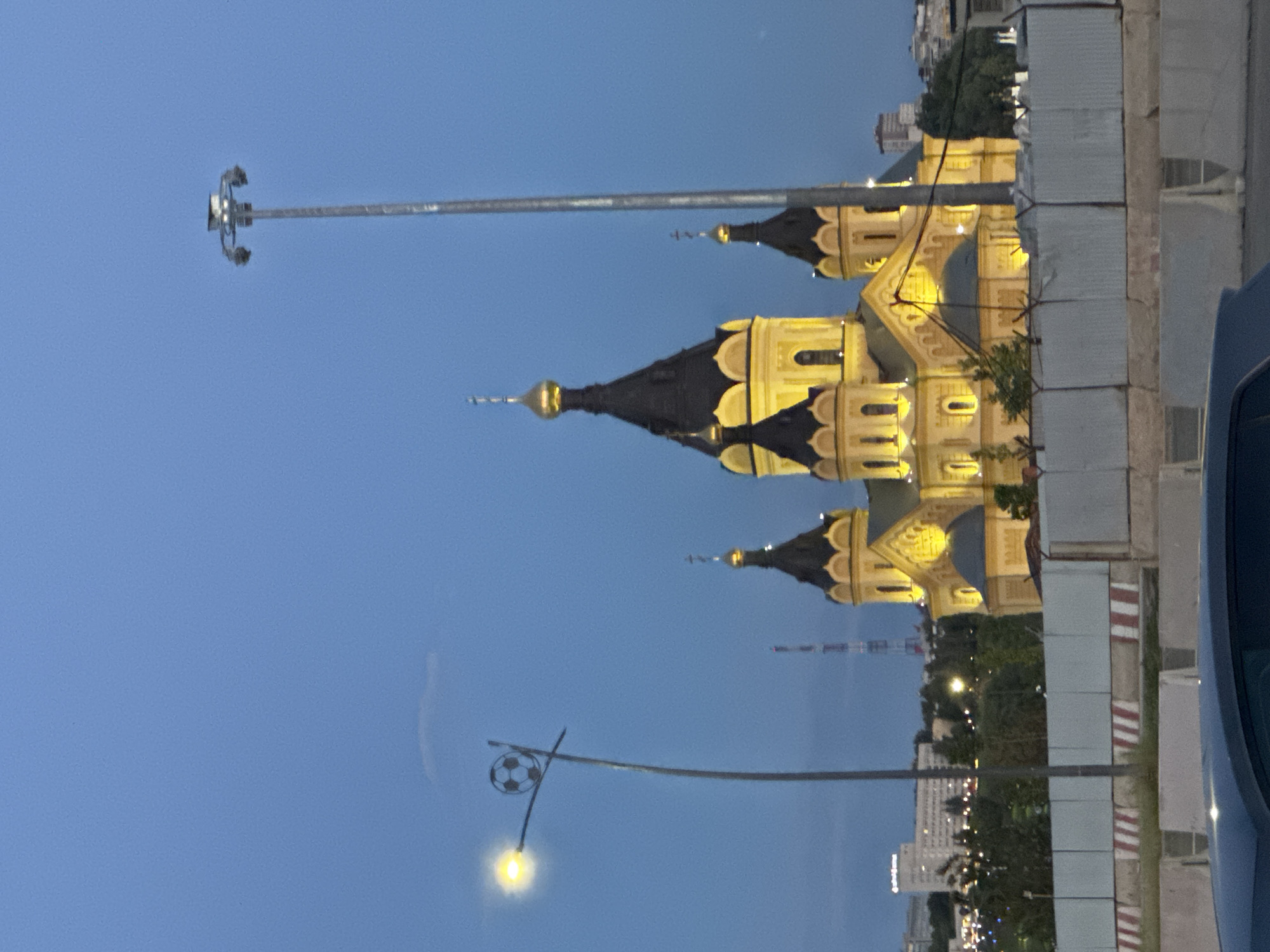 The image depicts an outdoor scene, likely in Nizhny Novgorod, Russia, during the twilight hours. The sky is a deep blue, indicating dusk or early evening. Dominating the central part of the frame are several golden-domed structures with dark roofs, characteristic of Orthodox churches. These buildings are illuminated from within, casting a warm glow that highlights their architectural details, such as arched windows and ornate spires topped with crosses.
In the foreground, a streetlamp with a distinctive soccer ball-shaped fixture is visible, emitting a soft yellow light. Another taller pole, possibly a light pole or part of a communication tower, extends from the left side of the image into the sky. To the right, a portion of a dark-colored vehicle is present, suggesting the image was taken from within or near a car.
The background reveals a cityscape with trees and buildings, some of which are illuminated by lights, further reinforcing the idea of a populated urban environment. The overall atmosphere is calm and serene, with the illuminated architecture providing a sense of grandeur against the darkening sky. There is no visible text in the image.