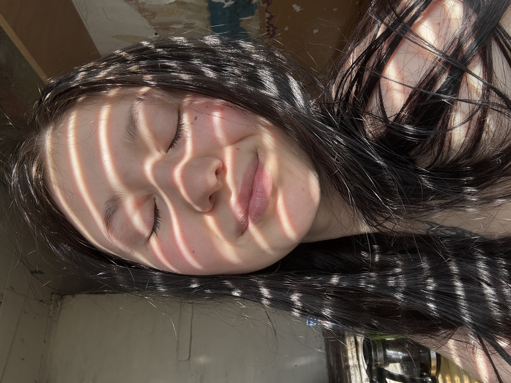 A young woman with dark hair lies with her eyes closed, possibly resting. Bands of sunlight stripe her face, indicating light filtering through blinds or a similar structure. Her hair is spread around her head. The background is indistinct but appears to be a wall with some discoloration or damage. The location in the city of Фоки, Russia cannot be determined from this image.