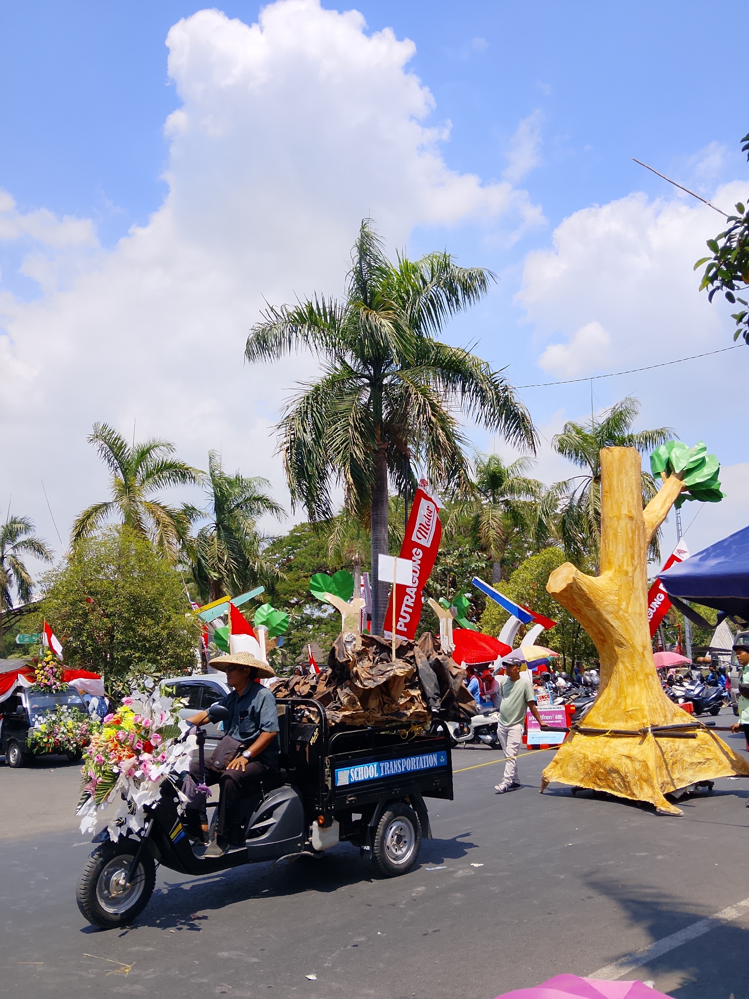 The image depicts an outdoor street scene, likely in Balun, Indonesia, given the context of the event it is part of. The sky is bright blue with scattered white clouds. In the foreground, a man wearing a straw hat and a dark shirt is seated on a three-wheeled vehicle, possibly an electric scooter. The vehicle is decorated with numerous colorful flowers, predominantly white and pink, with some yellow and orange accents. The vehicle's cargo bed is filled with what appears to be dried plant material, possibly representing branches or roots. The vehicle has "SCHOOL TRANSPORTATION" written in blue on its side.
To the right of the vehicle, a large, stylized yellow tree sculpture is prominently displayed, with a green canopy. Behind the sculpture, people are visible, as well as various flags and banners, some red and white, suggesting a festive or celebratory event. Palm trees and other lush green foliage are visible in the background, indicating a tropical or subtropical environment. To the left of the main vehicle, another vehicle, possibly a small truck, is also decorated with flowers. The ground is asphalt, and the scene appears to be a street or road. The overall atmosphere is bright and sunny.
