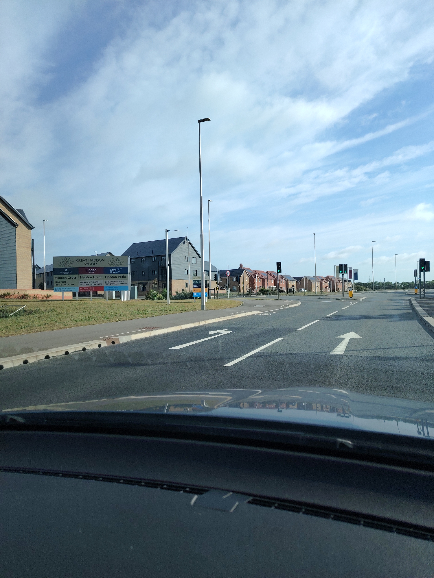 The image depicts a daytime scene from the interior of a vehicle, looking out through the windshield at a newly developed residential area and road intersection. The lower portion of the image shows the vehicle's dark dashboard and reflections on the windshield.
In the midground, a paved road features white lane markings, including a prominent straight arrow in the right-center lane and a left-turn arrow partially visible in the left-center lane. Multiple traffic lights are visible at the intersection, all displaying green signals. Streetlights with dark fixtures are positioned along the road, and a '30' mph speed limit sign is present on the left side. No pedestrians or other vehicles are clearly visible on the road or at the intersection.
To the left of the road, a large advertising sign for "GREAT HADDON WOOD" is displayed. The sign features logos for "Countryside," "Linden Homes," and "Bovis Homes," and details specific sections: "Haddon Cross" (listed as "SOLD OUT PHASE 2 DUE 2023"), "Haddon Green," and "Haddon Peake." Both "Haddon Green" and "Haddon Peake" mention "SHOW HOME IS OPEN 10AM-5PM SALES CENTRE" with associated phone numbers (01733 821 861 for Linden Homes, 01733 910 649 for Bovis Homes) and websites.
Behind the sign and along both sides of the road in the background are numerous modern residential buildings. These include multi-story apartment blocks with dark grey facades and roofs, and rows of individual houses with reddish-brown roofs. The architecture is consistent with recent construction. Grassy verges separate the roadway from these structures.
The sky is partially cloudy with patches of blue visible. The overall scene indicates a planned housing estate under development, located within the Great Haddon area, which is part of the administrative county of Cambridgeshire, United Kingdom. Huntingdonshire is a district within Cambridgeshire.
