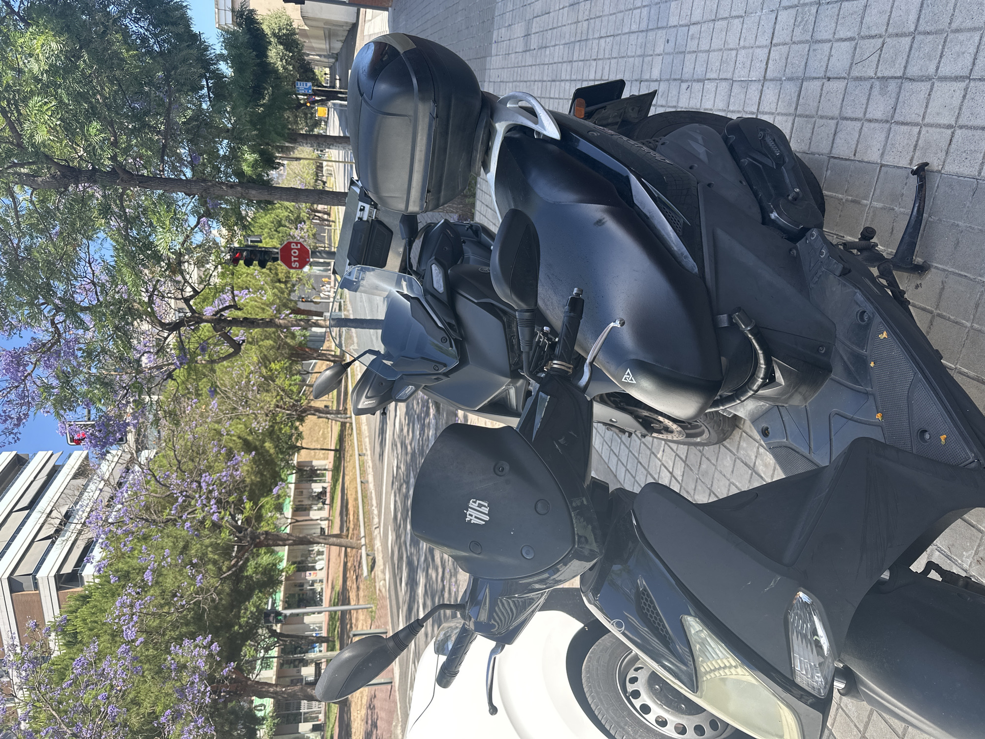 This image captures an urban street scene in La Fontsanta I Can Fatjó, Spain, on a clear, sunny day. In the immediate foreground, two black scooters are parked on a sidewalk composed of light grey square paving stones. The scooter closest to the viewer is black with a clear windscreen displaying a white, stylized "VANS" logo. It features a visible headlight, turn signals, handlebars with a chrome brake lever, and a black rearview mirror. Minor scratches are present on the front right fairing. A white car is partially visible to the left of this scooter. Behind the first scooter, a second dark-colored scooter is parked, equipped with a large black storage top case mounted on its rear rack. Its seat, a portion of its handlebars, and a black rearview mirror are visible.
In the midground, beyond the scooters and sidewalk, a paved street runs parallel to the foreground. A red traffic light with a single illuminated red signal is visible, accompanied by a circular red "STOP" sign on the same pole. Further down the street, another traffic light shows a green signal. The street is lined with numerous trees, many of which are adorned with vibrant purple blossoms, indicative of Jacaranda trees, interspersed with trees displaying green foliage. Across the street, a row of low-rise buildings houses ground-floor businesses with visible storefronts and awnings, topped by residential or commercial floors. Several indistinct human figures are visible walking on the distant sidewalk or near the street.
The background features a prominent, multi-story building with a light-colored facade and numerous windows, many equipped with dark, horizontal window blinds or awnings. The sky is clear and blue, occupying the upper portion of the frame. Additional trees and buildings are visible further in the distance under the bright daylight.