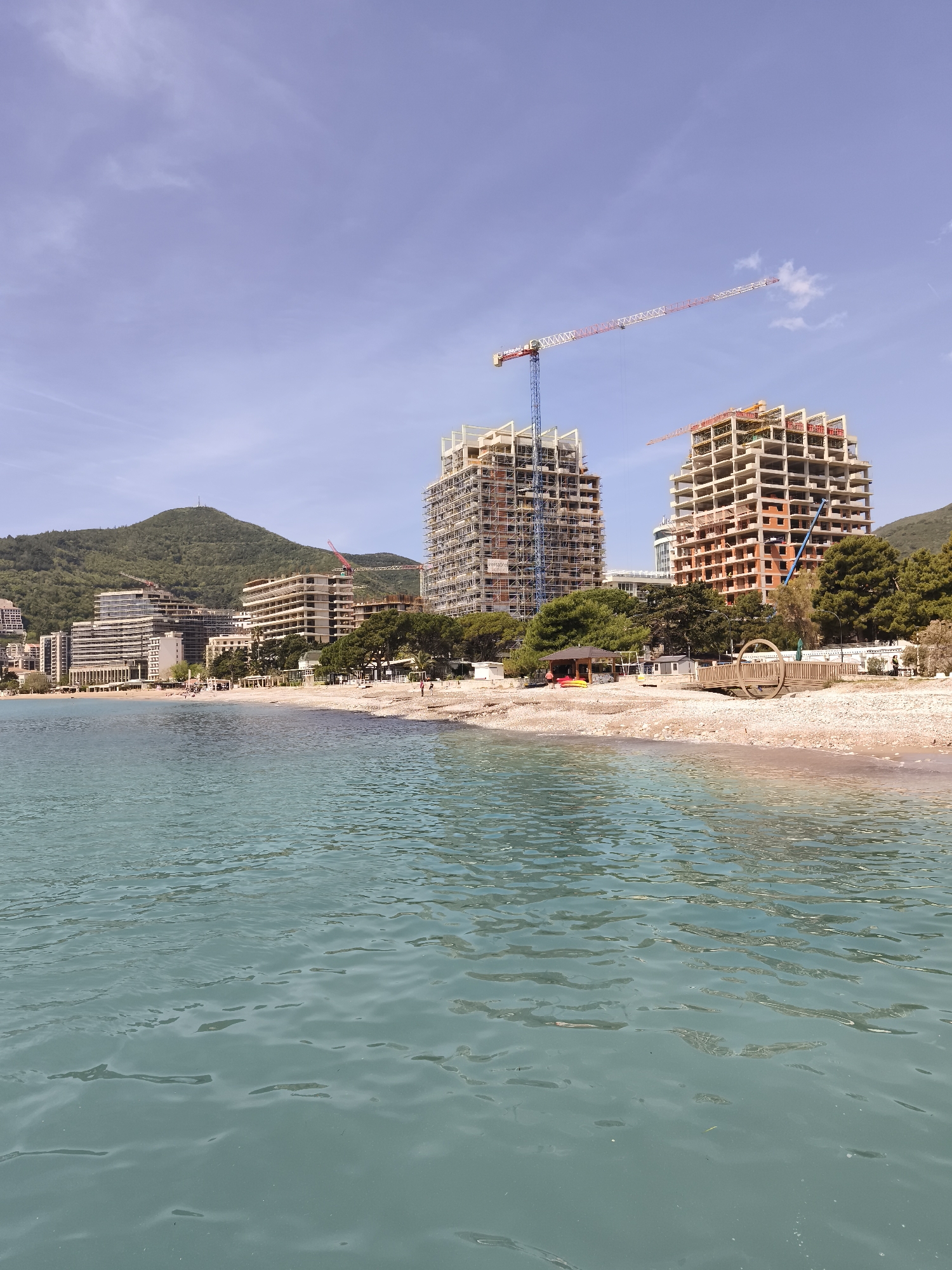 A medium, outdoor, eye-level shot shows a beach with water, buildings, and trees in Unknown, Unknown. The water is in the foreground and looks turquoise blue. The beach is a light brown color. There are a few wooden structures on the beach as well as trees. There are tall buildings beyond the beach with scaffolding and cranes around them. Behind the buildings is a green hill and a light blue sky.
