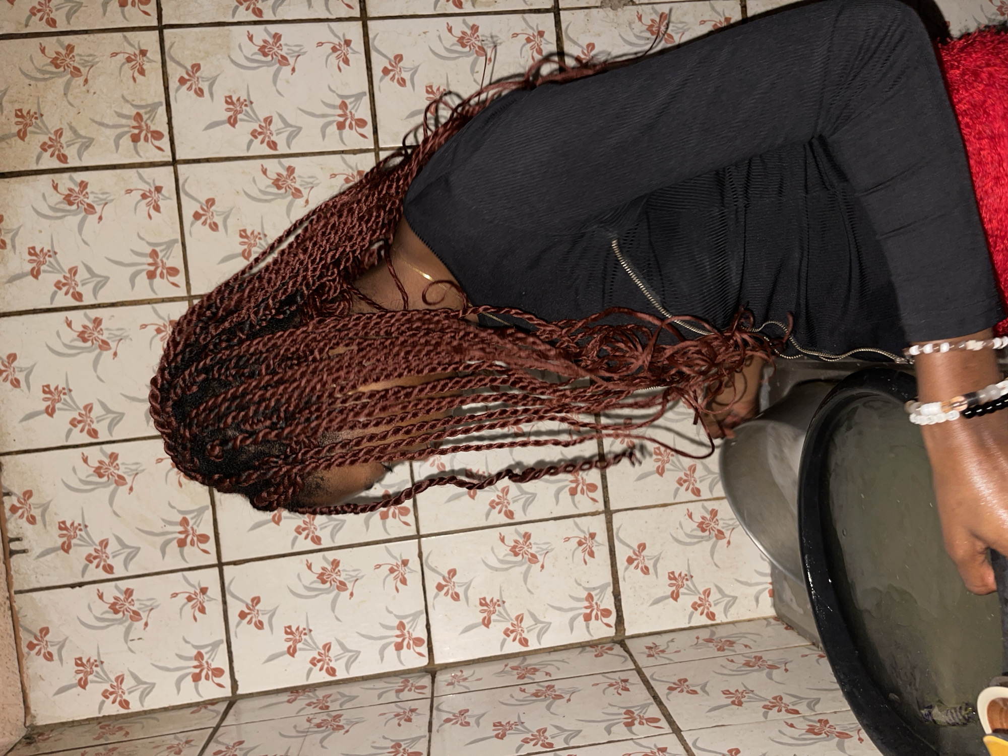 The image shows a person with reddish-brown braided hair leaning over a toilet. The person is wearing a black long-sleeved shirt and bracelets on their wrist. The toilet appears to be in use. The floor is tiled with patterned tiles. The location is Unguwar Gabas, Nigeria.
