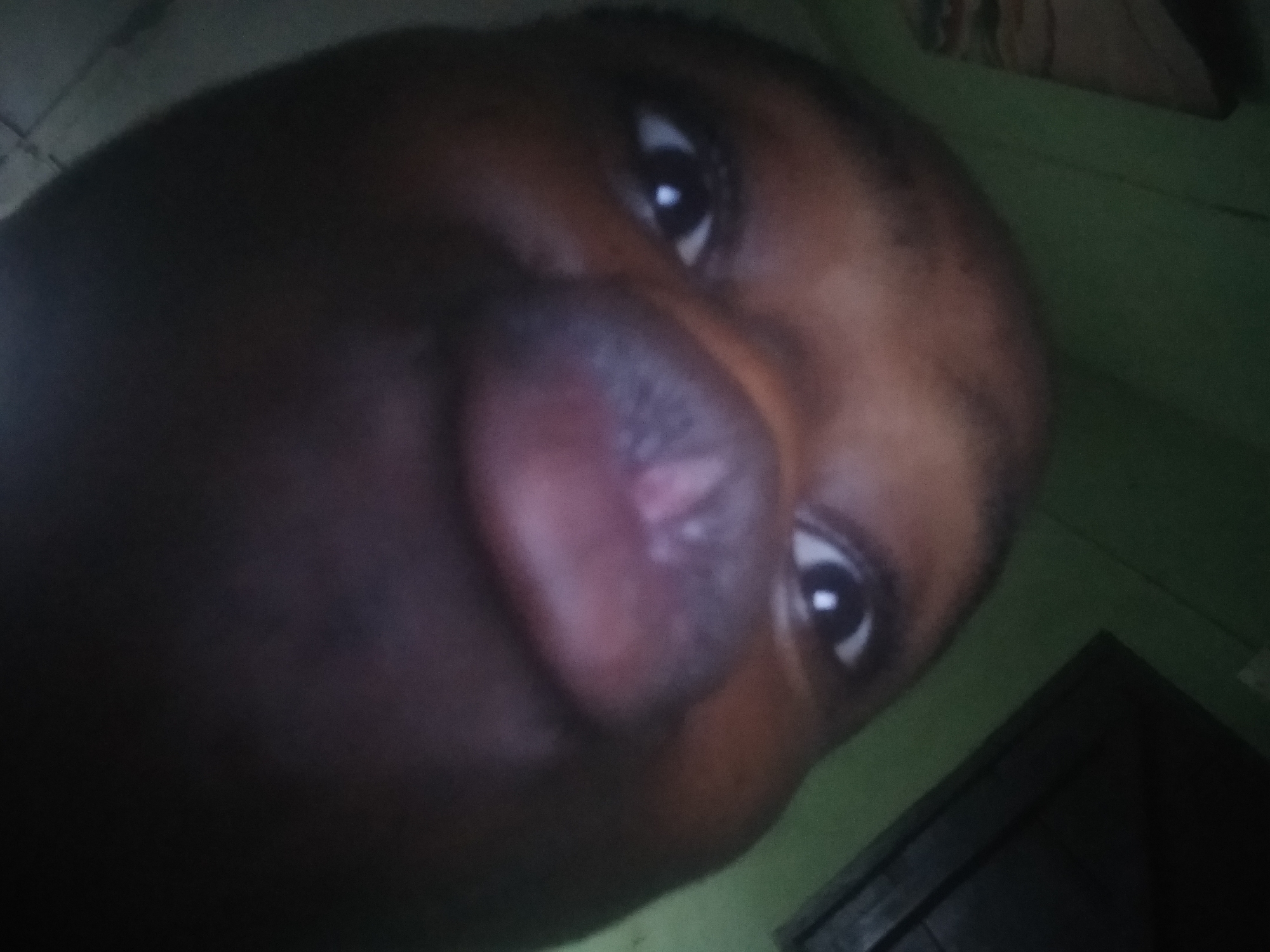A close-up, slightly tilted image shows a person with dark skin. The focus is on their face, particularly their lips, nose, and eyes. The lighting is dim, casting shadows across the face. A wall with green color is behind the person. A dark corner of the wall, likely a door or cupboard, is slightly visible on the right side. The location of the scene is Shangisha, Nigeria.