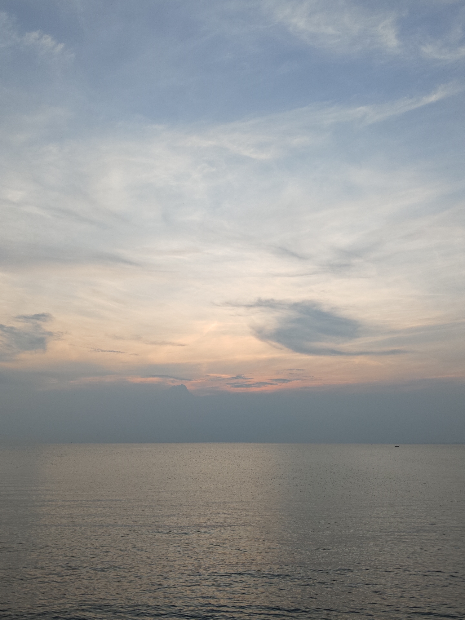 The image shows a seascape at dusk or dawn. The sky is layered with shades of blue and streaks of pink and orange. A band of dark gray clouds hovers above the horizon. The water is calm with gentle ripples, reflecting the colors of the sky. A small boat is visible on the horizon. The location is Lhokseumawe, Indonesia.