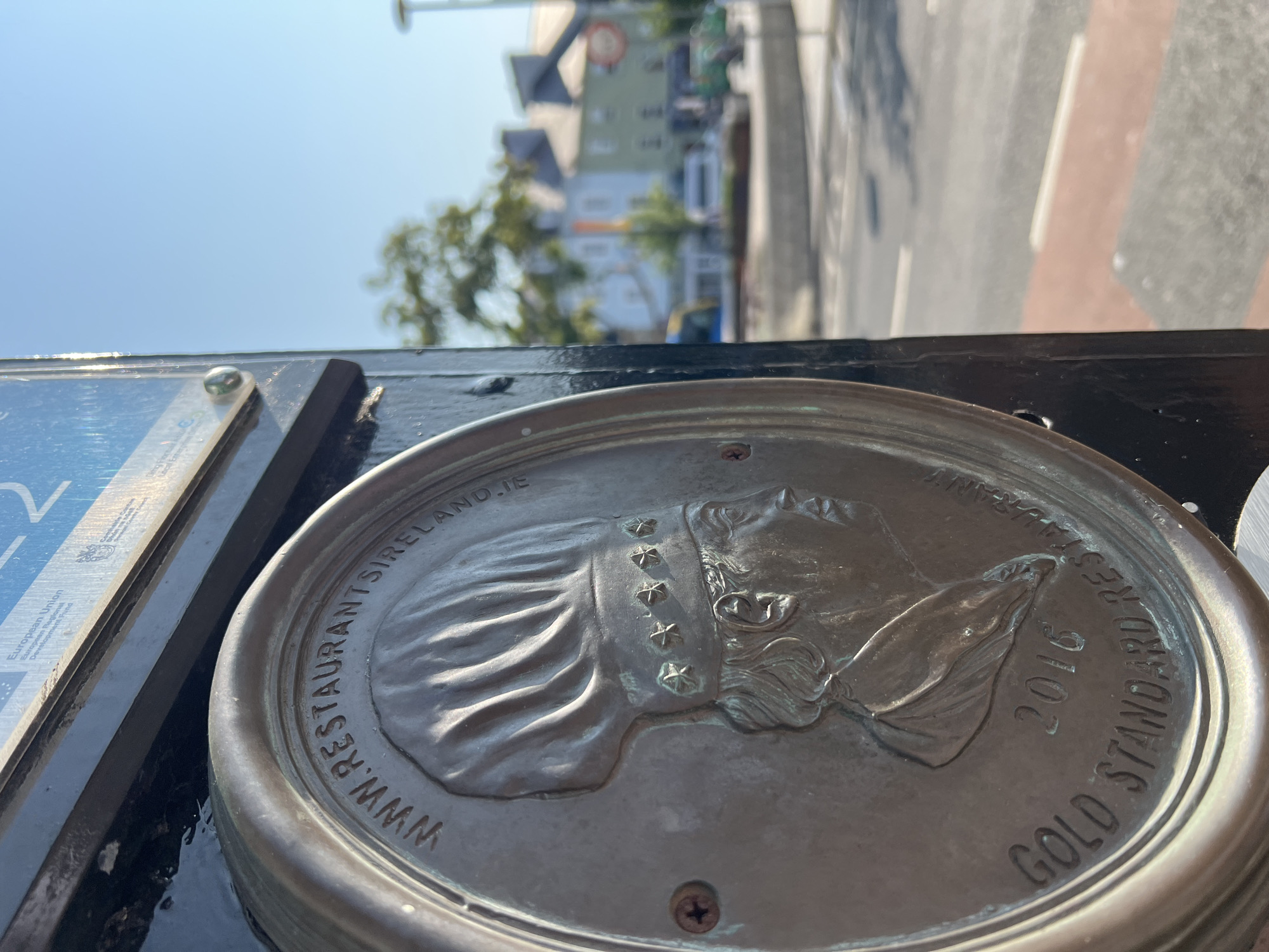 A close-up, eye-level, outdoor shot focuses on a bronze plaque affixed to a dark, rectangular post. The plaque is oval-shaped and features a relief of a chef's profile wearing a tall hat adorned with stars. Inscribed on the plaque are the words "WWW.RESTAURANTS IRELAND.IE" along the top curve and "GOLD STANDARD RESTAURANT 2016" along the bottom curve. A single screw is visible to the left of the chef's profile. The background, out of focus, shows a street scene with buildings, trees, and a clear blue sky. This is a close-up view of an award plaque for a restaurant located in the city of Donegal, Ireland.