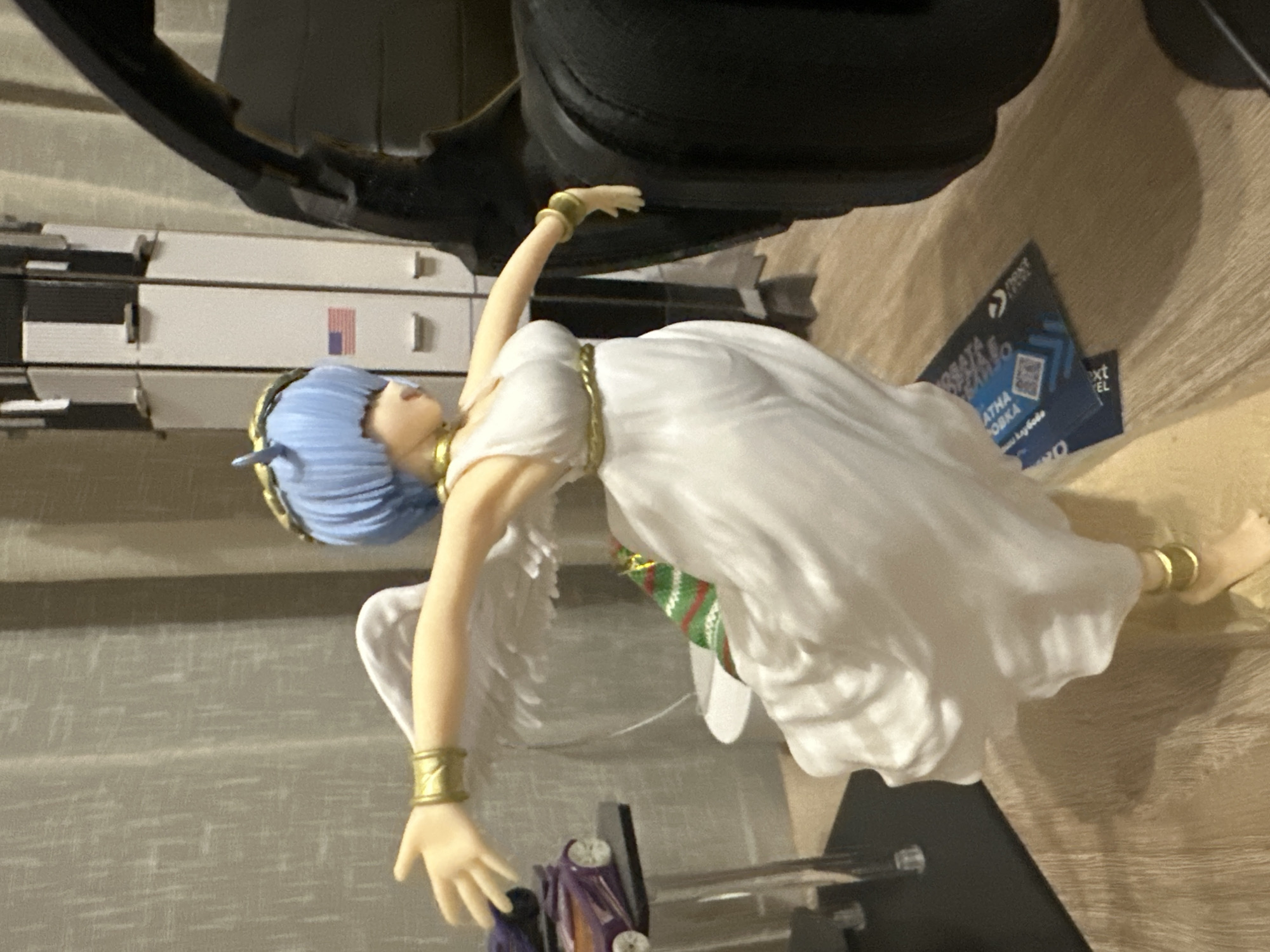 An anime-style figurine of a woman with blue hair, white wings, and a white dress is displayed in what appears to be a room. She wears a gold headband and gold armbands. Behind her, a model of a rocket is visible, featuring an American flag decal. To the left, a toy car sits on a clear acrylic stand. A black object, possibly headphones, is positioned to the right of the figurine. The floor is made of wood-like material, and a patterned curtain is in the background.