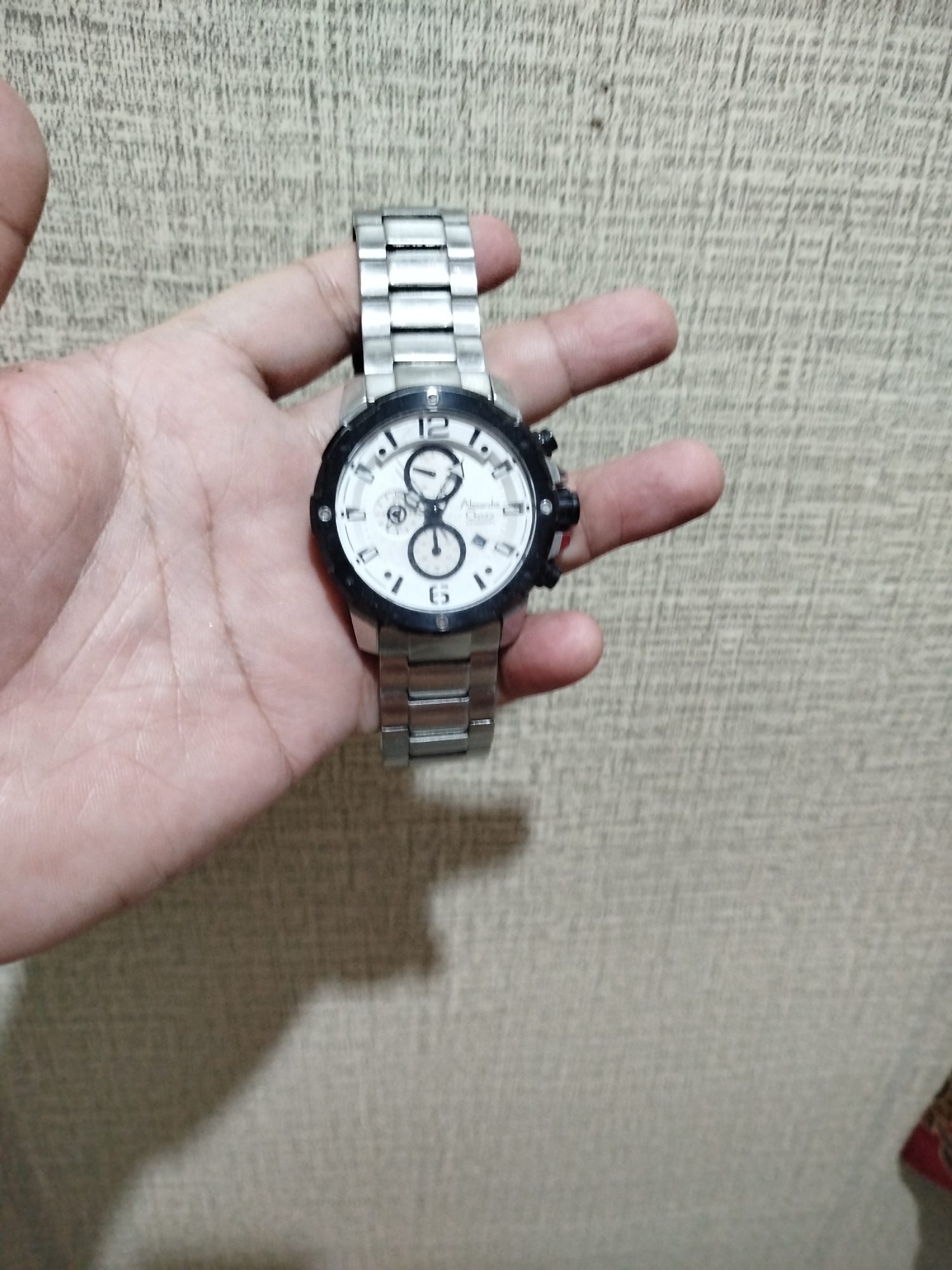 A wrist watch with a white face and metal band is held in a person's left hand. The watch has three subdials and a date window. The background is a textured, light brown wall. This image was taken in Bandungrejo, Indonesia.