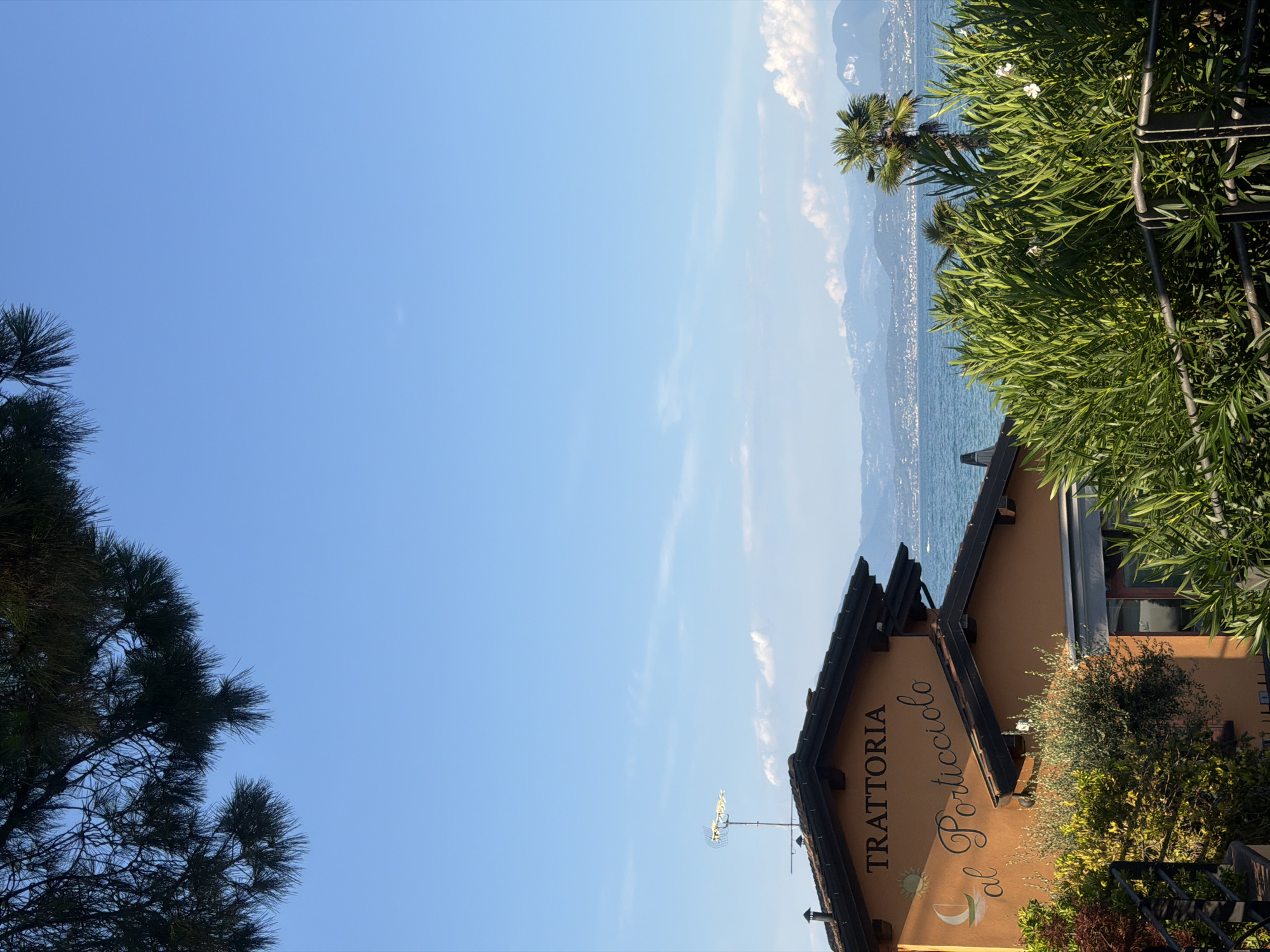 The image shows a bright, sunny day in Sirmione, Italy, with a clear blue sky and a few wispy clouds. In the foreground, lush green foliage obscures a metal railing. Behind the foliage, a building with an orange facade is visible. The building has a dark tiled roof and an antenna on top. Text on the facade reads "TRATTORIA al Porticciolo", indicating it is a restaurant. A small illustration of a sailboat and a sun is also present on the facade. Beyond the building, a body of water, likely Lake Garda, stretches out, with a distant shoreline featuring buildings and mountains. A palm tree stands tall to the right of the building, adding to the Mediterranean atmosphere. The scene is peaceful and serene, evoking a sense of vacation and relaxation.