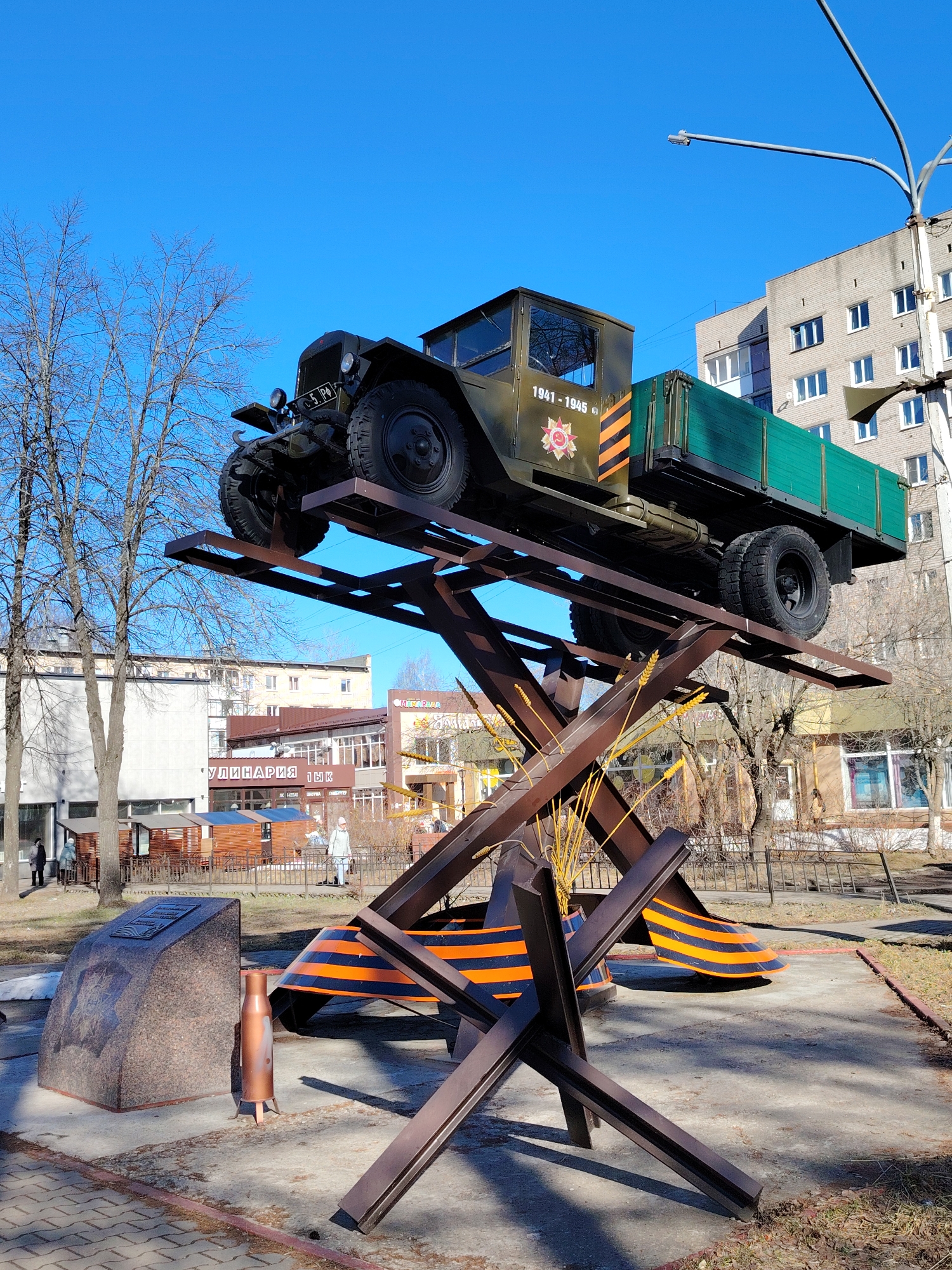 The image shows a military vehicle mounted on a metal structure in the city of Chaikovsky, Russia. The vehicle, identified as a GAZ-AA truck, is painted in military green and features markings that indicate it served in World War II (1941-1945). The truck is elevated on a dark brown metal framework designed to resemble anti-tank obstacles. Orange and black striped banners, known as the Ribbon of Saint George, are incorporated into the structure. A granite stone with a commemorative plaque and a large caliber shell casing stand nearby. In the background, several multi-story buildings and bare trees are visible under a clear, blue sky. Pedestrians are seen walking in the background. The scene appears to be a war memorial or monument within a public park or plaza.
