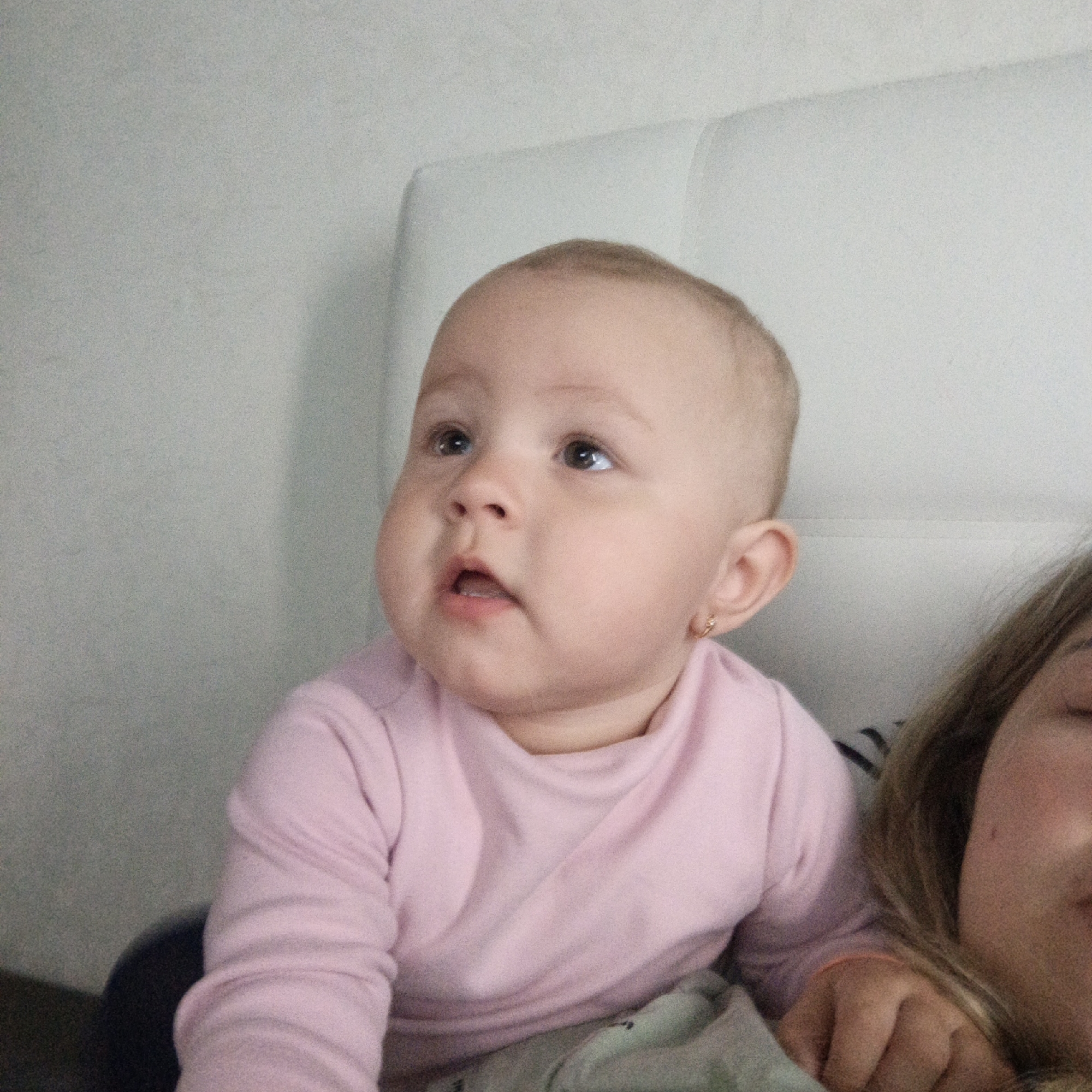 The image shows a baby girl with fair skin and light brown hair wearing a pink long-sleeved shirt. She is looking upwards with her mouth slightly open. She has an earring in her left ear. The baby is sitting in front of a white headboard. A person with light brown hair is partially visible on the right side of the image. The location is Unknown, Unknown.