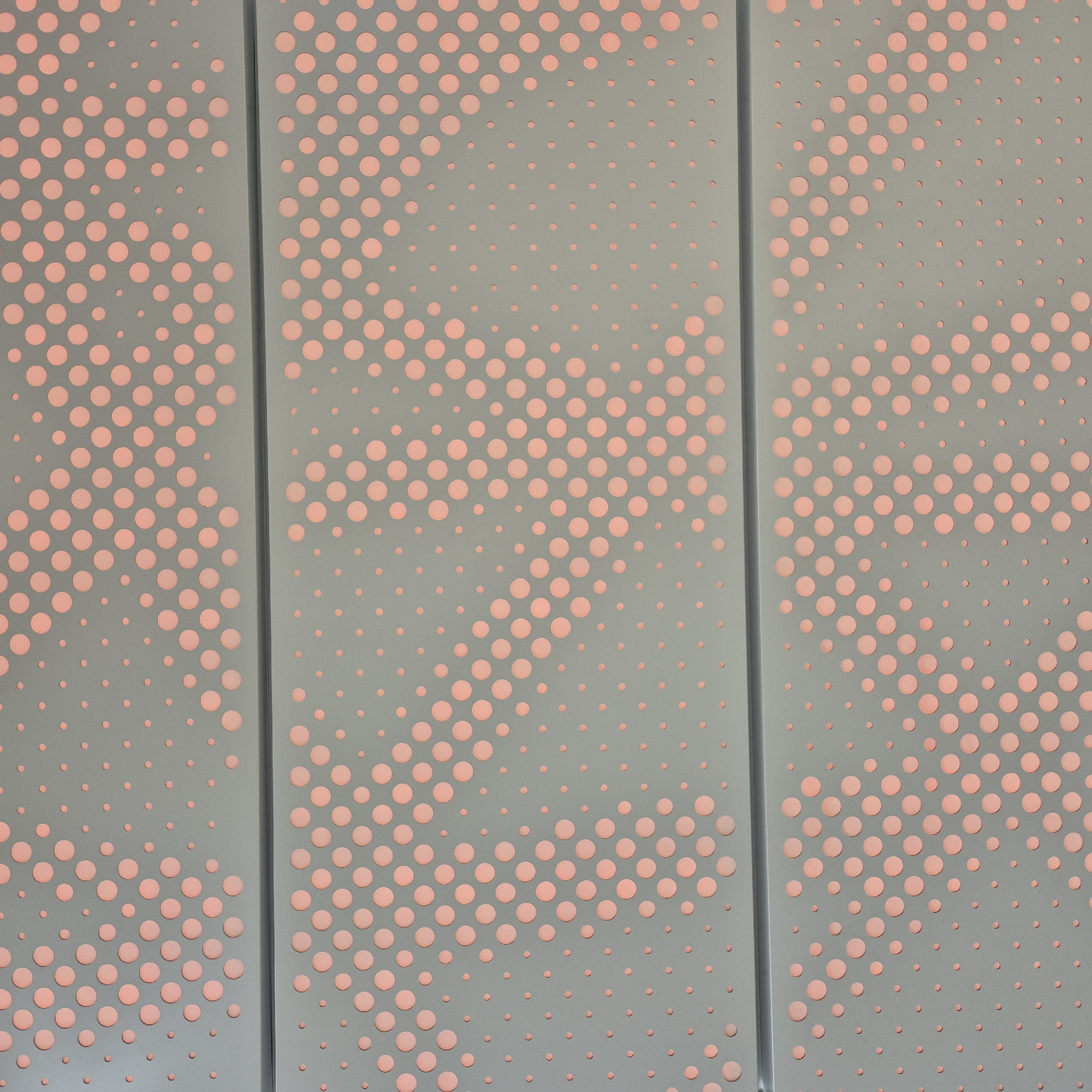 The image shows a close-up of a gray perforated panel with a pinkish-orange backing. The perforations are circular and vary in size, creating a gradient effect of dots. The panel is divided into three horizontal sections by thin, dark gray lines, suggesting it is part of a larger structure, possibly a wall or ceiling. The dots are denser and larger in some areas, transitioning to smaller and sparser dots in others, forming a pattern that is reminiscent of a halftone print. The lighting appears to be from behind the panel, illuminating the dots and creating a soft glow. There are no people or any other discernible objects in the image. The scene is abstract and focuses entirely on the texture and pattern of the perforated material.