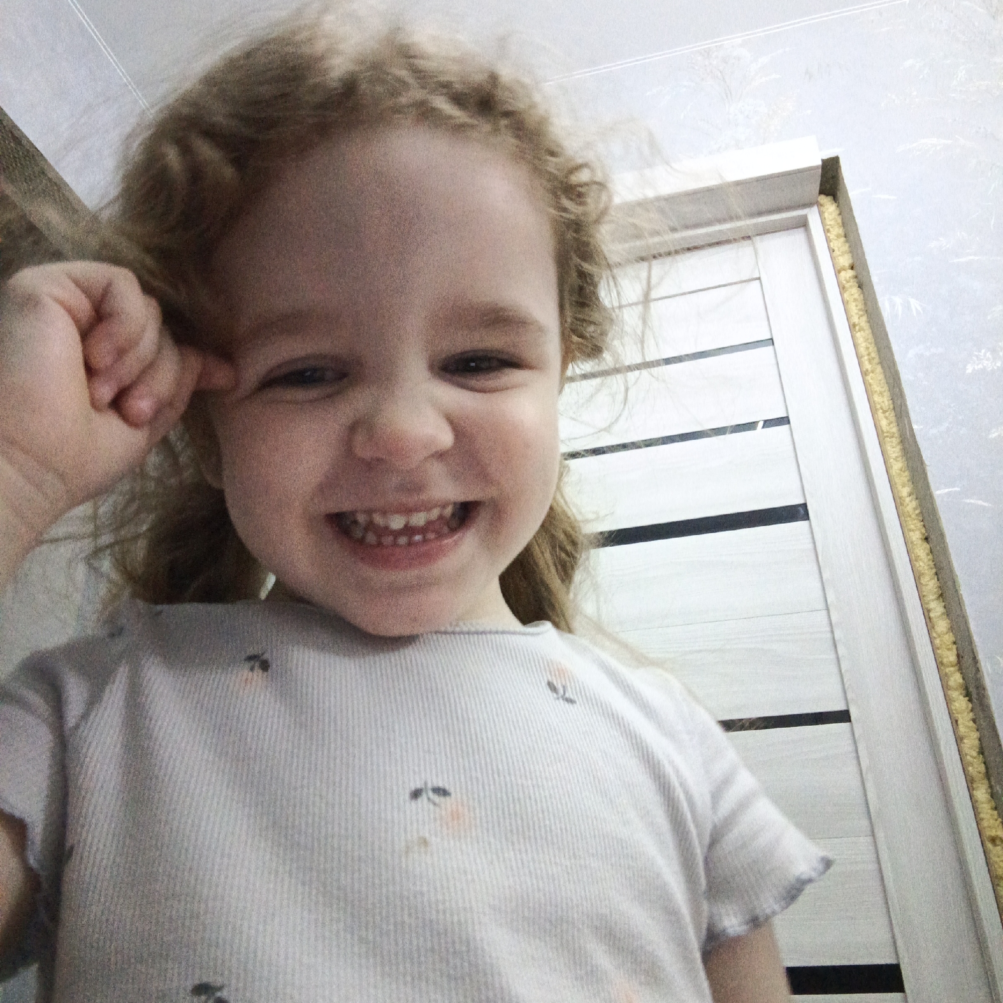 A joyful young child with light, curly hair is captured in a close-up, upward-angled shot. The child, likely a toddler or preschooler, is smiling widely at the camera, revealing small teeth, and has dark eyes. One hand is raised, touching the side of their head near their temple. They are wearing a light-colored, ribbed t-shirt with small, dark patterns resembling insects and some lighter orange/pink smudges on the fabric.
The background features a white door with horizontal dark lines or grooves, positioned to the right of the child. To the right edge of the doorframe, yellow expanding foam insulation is visible, suggesting ongoing construction or renovation of the doorway. The adjacent wall is covered with light-colored wallpaper featuring a subtle, faint pattern, possibly floral or organic.
The scene is indoors, likely a home in Марковский, Russia. The child's expression conveys happiness and playfulness. No visible text is present in the image.