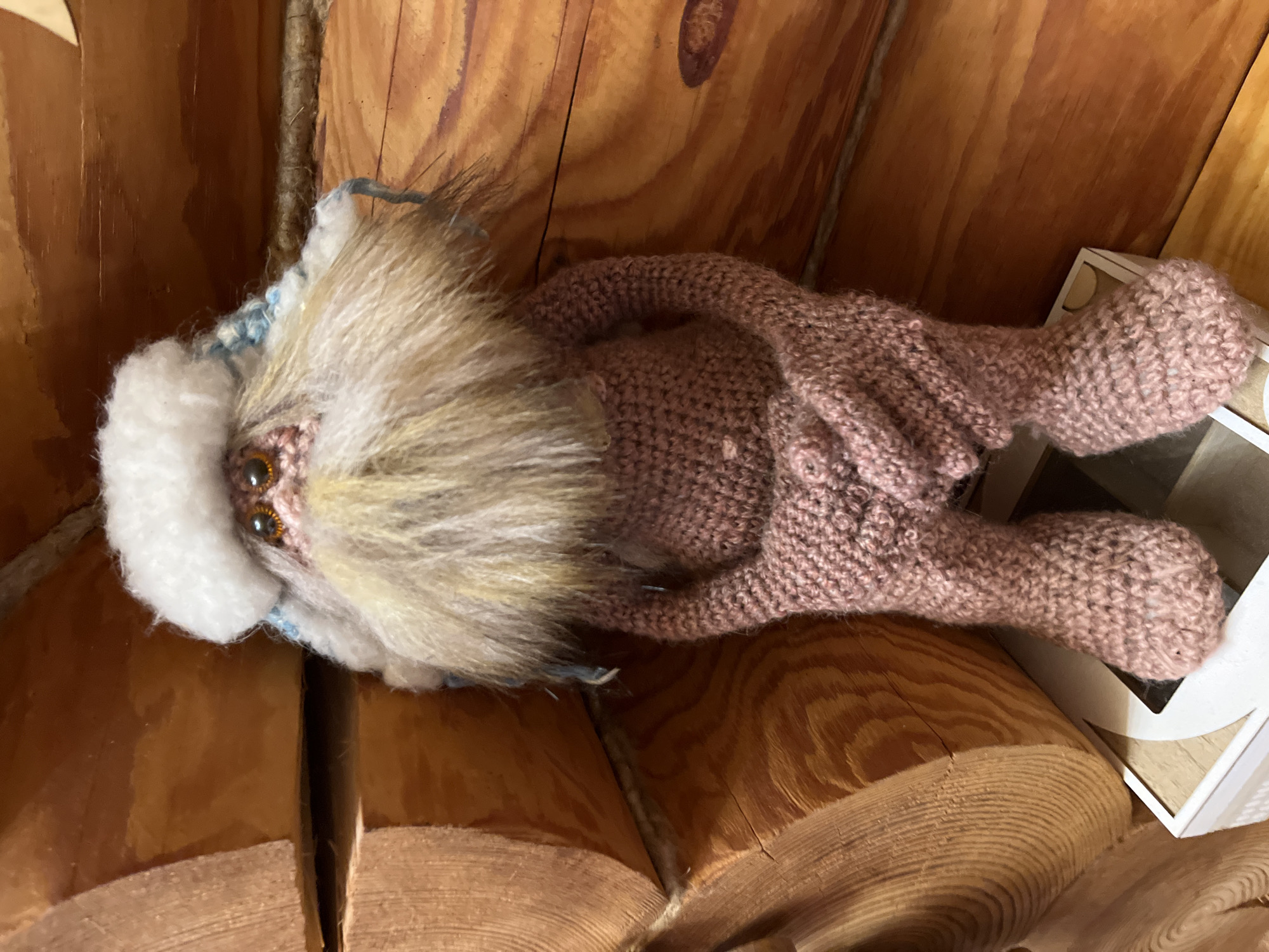 A handcrafted, plush doll resembling a mystical creature lies horizontally across a surface constructed of light-brown, rounded wooden planks. The doll has a head with a white fur-trimmed hood, a tan fur-like beard, and amber-colored beaded eyes. The doll's body is crocheted in a dusty-rose color and appears to be loosely shaped, with attached limbs positioned in a reclining posture. Part of a white box is visible near the doll's feet. The scene suggests an interior setting with a focus on handcrafted items and wooden construction. The location is Chaikovsky, Russia.