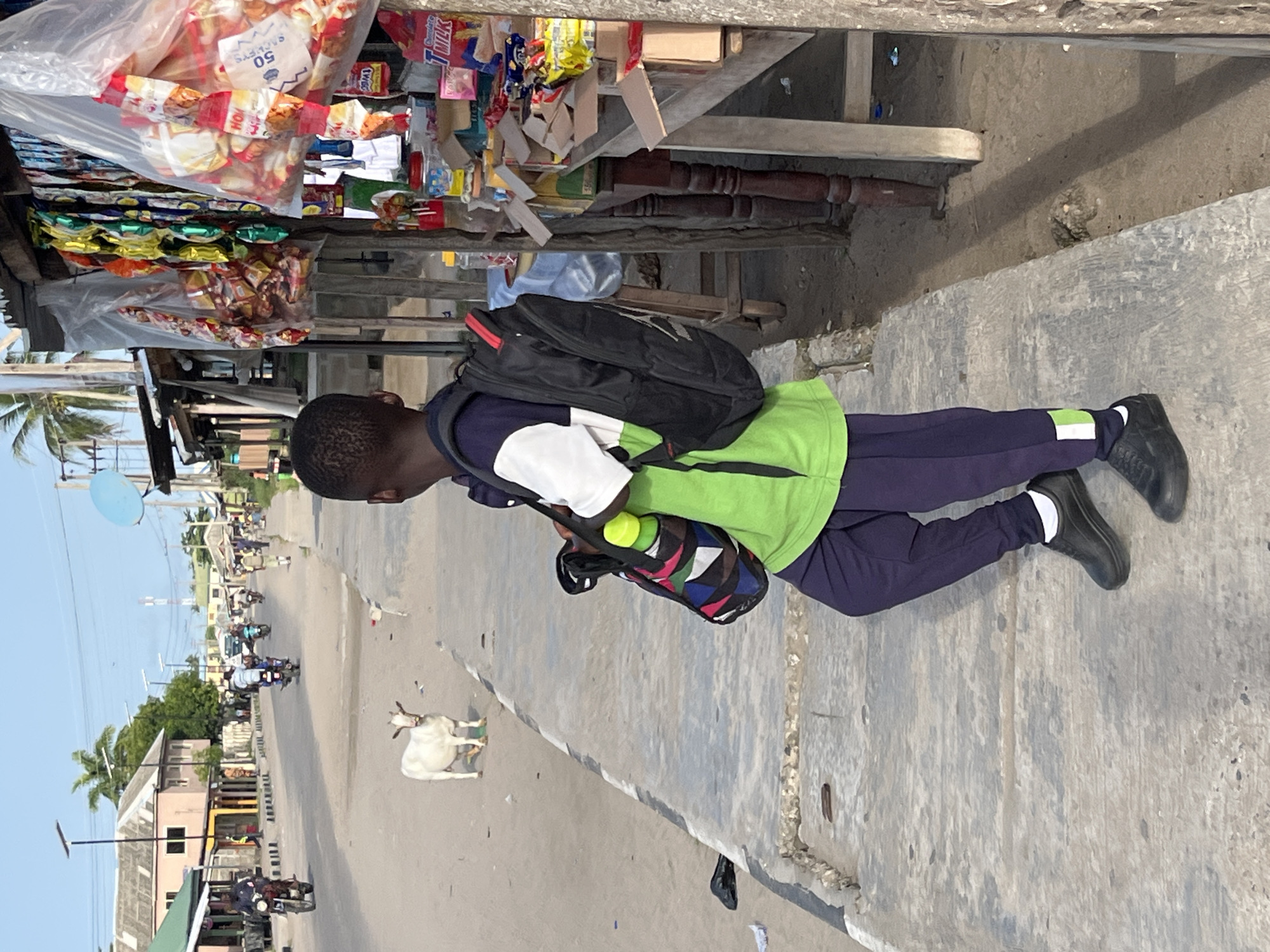 A young person walks on a concrete sidewalk in Badagry, Nigeria. A small roadside shop selling snacks and candy is on the left. The individual is wearing a black backpack, a green shirt, and dark pants with reflective stripes at the ankles. They are also carrying a colorful patterned bag and a green water bottle. In the street, there is a white goat and several motorbikes. Buildings and trees are visible in the background.