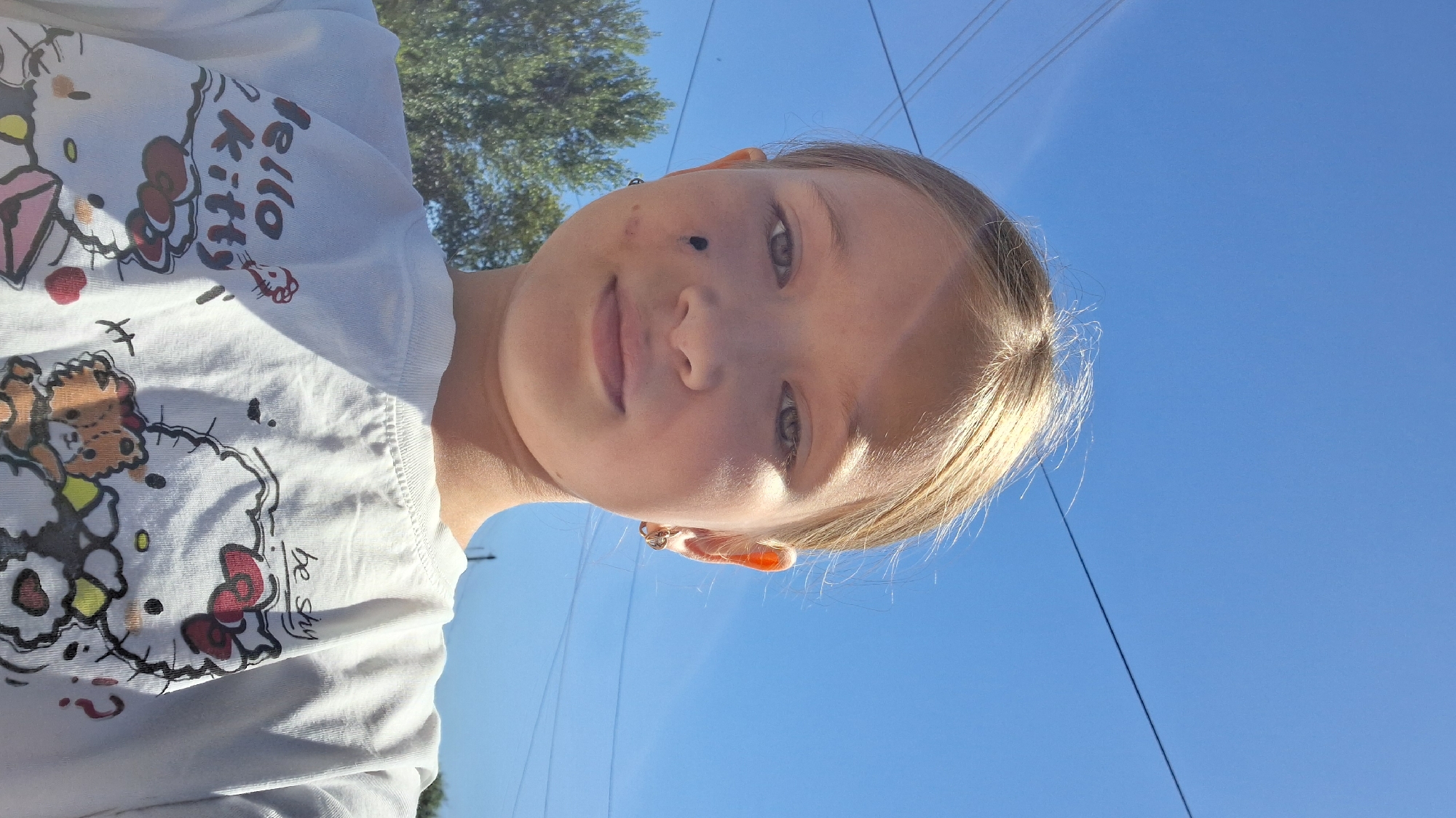 The media file is a portrait-oriented, close-up photograph of a young girl outdoors, looking at the camera. The girl has fair skin, blonde hair, and light-colored eyes. She is wearing a white t-shirt with a graphic print featuring Hello Kitty characters and the text "Hello Kitty". A small, golden hoop earring is visible in her right ear.
The background consists of a clear, bright blue sky, suggesting a sunny day. The sun is casting a warm light on the girl's face, creating highlights on her hair and cheek. The top portion of a green tree is visible in the upper left corner. In the distance, thin power lines stretch across the sky.
The photograph appears to be a selfie, as indicated by the angle and the close proximity of the subject. The girl's expression is neutral, and her gaze is directed towards the viewer. The overall impression is one of a casual outdoor moment on a bright, clear day, likely in Semey, Kazakhstan, given the provided location context. There are no discernible emotions conveyed beyond a general sense of being present.