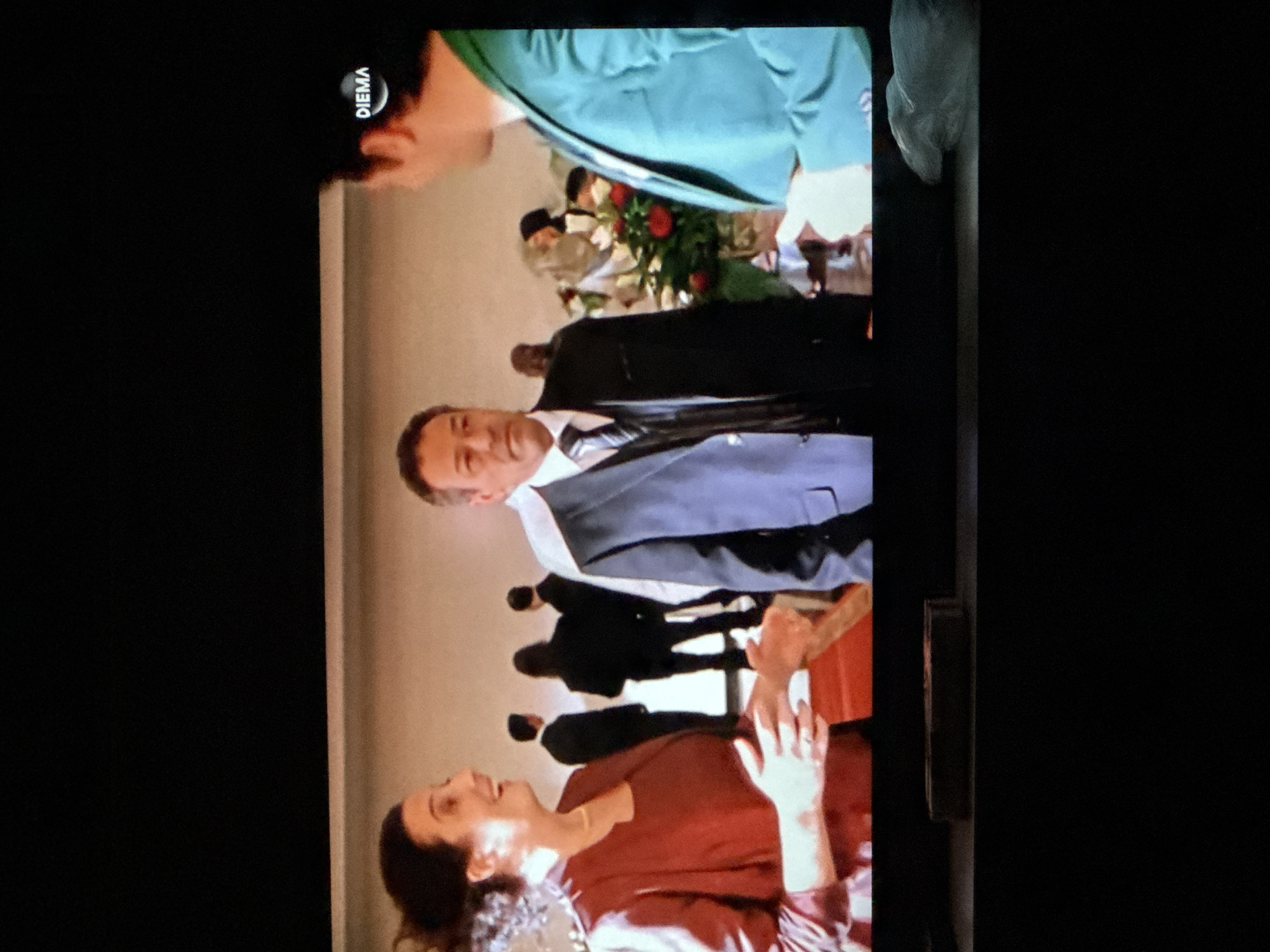 A low-angle shot of a television displaying a scene with multiple people, possibly filmed in Sarnevo, Bulgaria, though this is not directly visible in the image. A woman in a red shirt is at the bottom of the frame, smiling and holding her hands up. Above her is a man in a grey suit with a black tie. Behind him is a group of people dressed in black suits. In the background, there is a display of red roses and greenery. A person in a green shirt is in the top of the frame. The logo for the television channel Diema is in the top left corner of the frame.