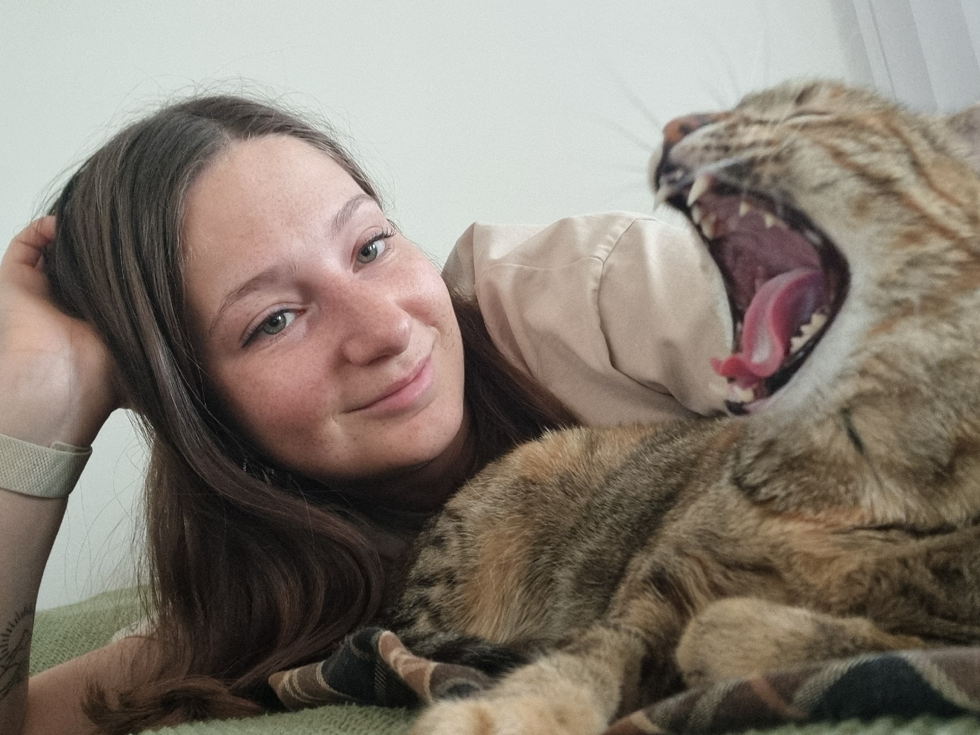 A woman is lying down next to a cat. The woman has long brown hair, fair skin, freckles, and is smiling. She is wearing a light-colored shirt and a watch. The cat is lying on a checkered blanket and is in the process of yawning, revealing its teeth and tongue. The background is a plain light color. No information suggests that the image was taken in Ramat Gan, Israel.