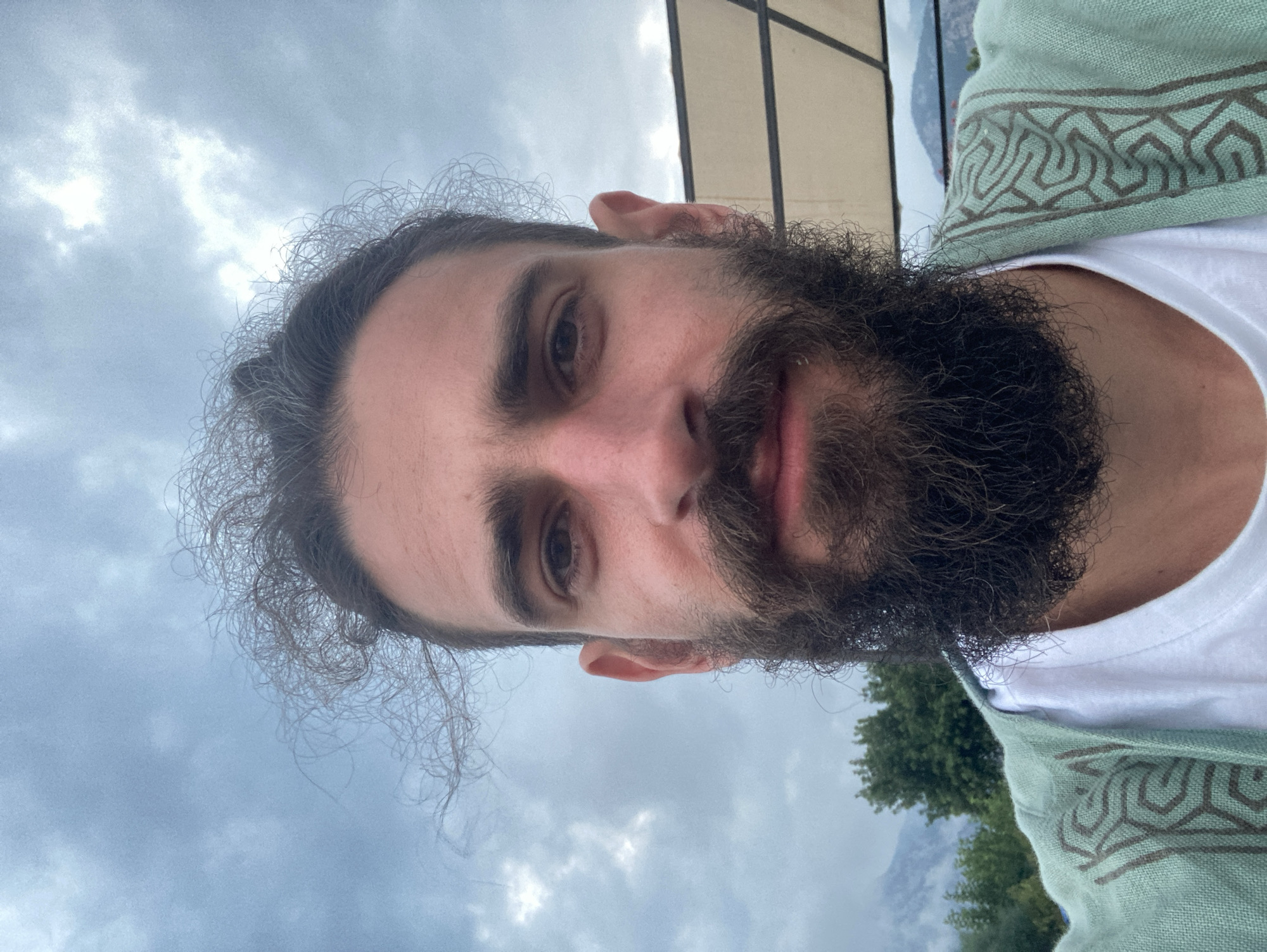 A close-up, front-facing selfie features a person with a dark, full beard and long, dark, curly hair pulled back. They are wearing a white t-shirt and a light green, patterned garment over it. The background consists of a cloudy sky, trees, and mountains, suggesting an outdoor setting, possibly in or around Kemer, Turkey. A portion of a tan awning or structure is visible in the upper right corner.