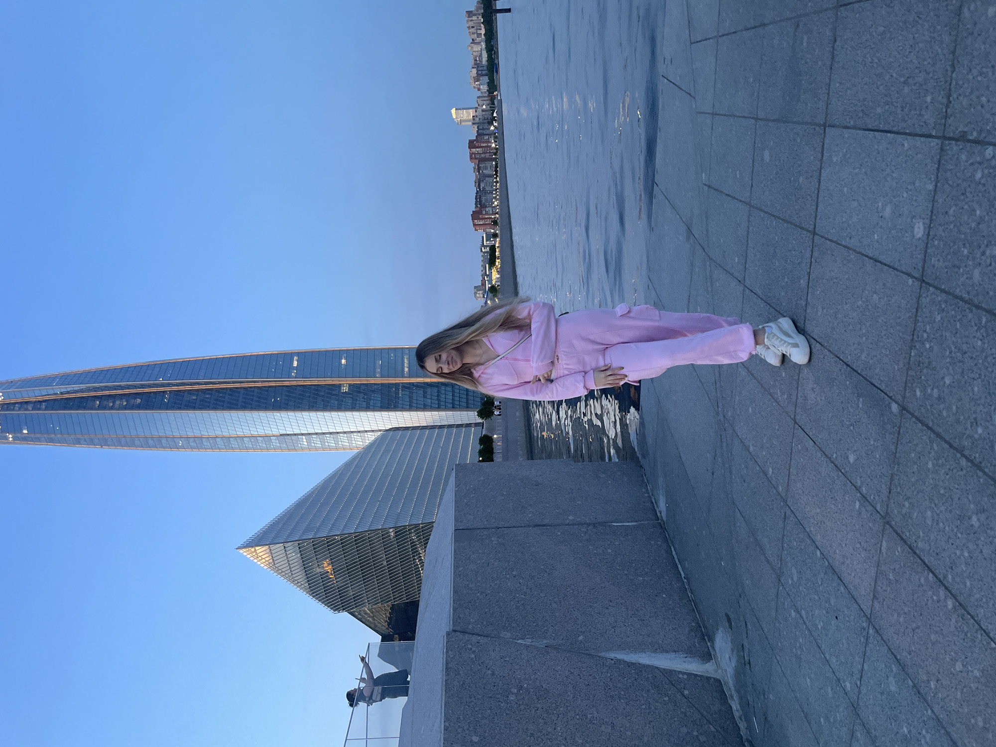 A young woman with blonde hair is posing for a photo outdoors at dusk in front of a body of water, with a modern cityscape in the background. She is wearing a pink hooded jumpsuit with cargo pockets and white sneakers. She stands on a paved walkway, with her left arm casually resting on her hip. Behind her to the left, a triangular glass building and a tall, slender skyscraper are visible. In the distance, across the water, is a line of buildings. A second, indistinct figure is visible on a railing to the left of the frame, looking out towards the water and pointing. The sky is a clear blue.