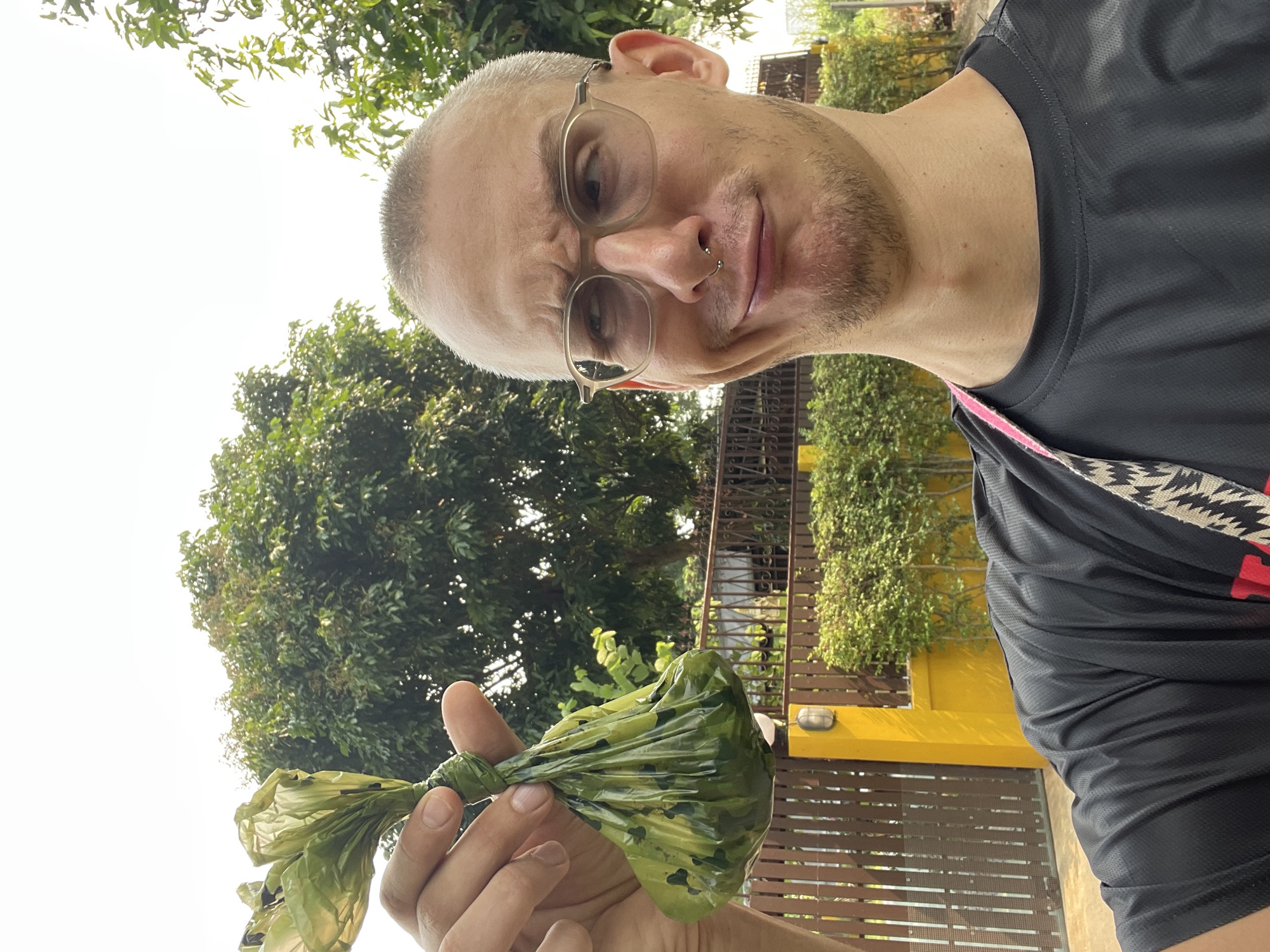 A man takes a selfie outdoors, holding up a twisted green plastic bag, likely containing dog waste. He has short, light-colored hair, a light beard, and wears glasses with light frames and a small gold-colored nose ring. His facial expression is a slight grimace, suggesting mild annoyance or exasperation. He is wearing a dark, possibly black, short-sleeved athletic shirt, with a patterned shoulder strap visible over his right shoulder.
The background is filled with lush green foliage and trees under a bright, white sky, indicating daylight with potential haze or overcast conditions. In the midground, there's a structure with horizontal slats and sections painted bright yellow, possibly part of a building or fence. The setting appears to be a residential or public outdoor area in Nong Pa Khrang, Thailand. No visible text is present.