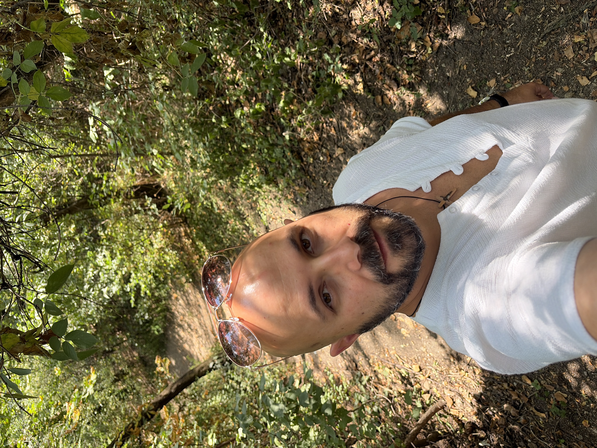 A man with a shaved head and a beard is taking a selfie outdoors in a wooded area. He is wearing a white shirt with a V-neck and buttons, and sunglasses are perched on his head. The background is filled with green foliage and a path winding through the trees. The lighting suggests it is daytime, with sunlight filtering through the leaves. The location is identified as the Vitosha mountain near Sofia, Bulgaria.