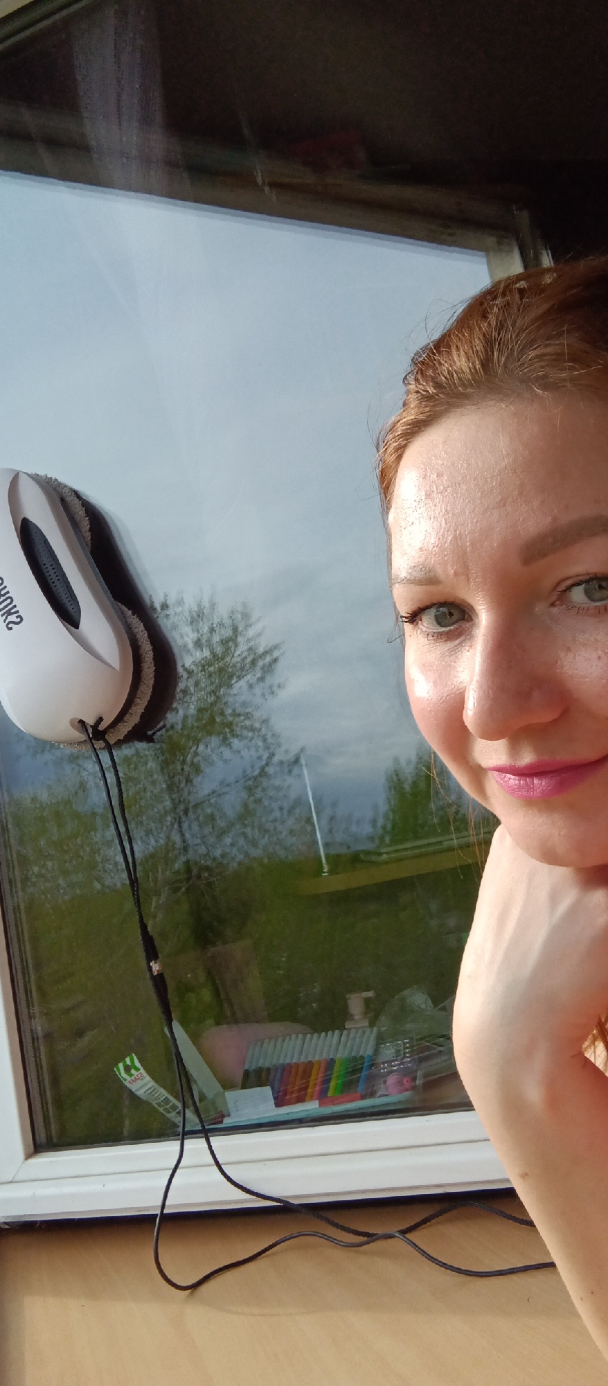 A woman is positioned close to a window, capturing a selfie. The window features a white automatic glass cleaning tool attached to it. The outside environment reflecting on the window shows greenery and a partly cloudy sky. Inside, on the window sill, there are several colored markers or pens, along with some other items, possibly cleaning or household products. The woman has her head resting on her hand, and she is smiling slightly at the camera. Her hair is reddish-brown, and she is wearing lipstick. The setting seems to be indoors, possibly in a home.