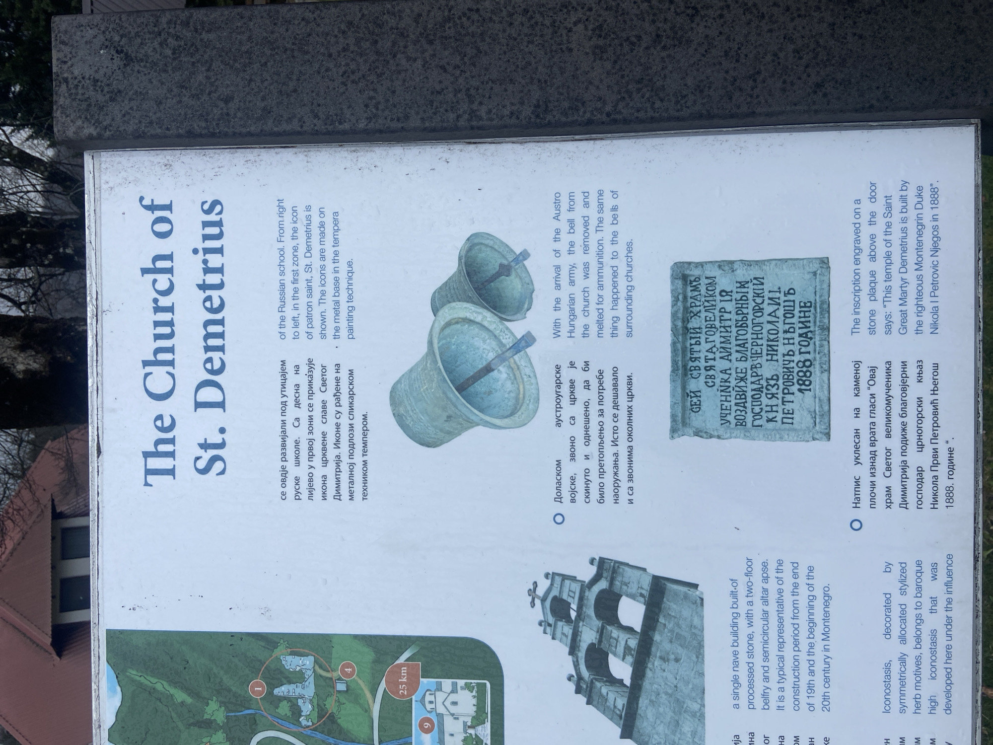 The image is a signpost for the Church of St. Demetrius in Kolašin, Montenegro. The sign is mounted on a dark gray stone base. It provides information about the church in both Serbian and English, including details about its history, architecture, and artistic elements. A map shows the location of the church. The sign includes images of the church's bells and an inscription plaque on the building. The information on the sign covers topics such as the influence of the Russian school, the removal of bells by the Austro-Hungarian army, and the inscription about the building's construction by Duke Nikola I Petrovic Njegos in 1888.
