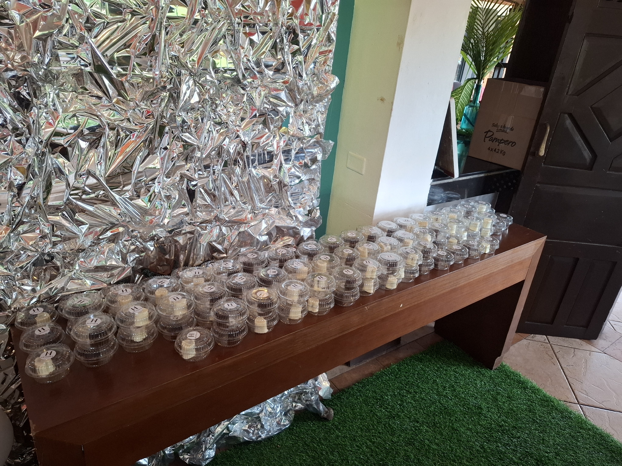 The scene depicts a long, dark wooden table covered with numerous clear plastic containers of individual desserts, possibly in Playa El Agua, Venezuela. The containers are arranged in neat rows, each bearing a small sticker with the letter "H". Behind the table, crumpled silver foil covers a wall. To the right, a beige wall and a partially visible box labeled "Pampero 4x4,2Kg" rest next to an open, dark wooden door. The floor is tiled in the bottom right and is covered with a patch of artificial green grass.