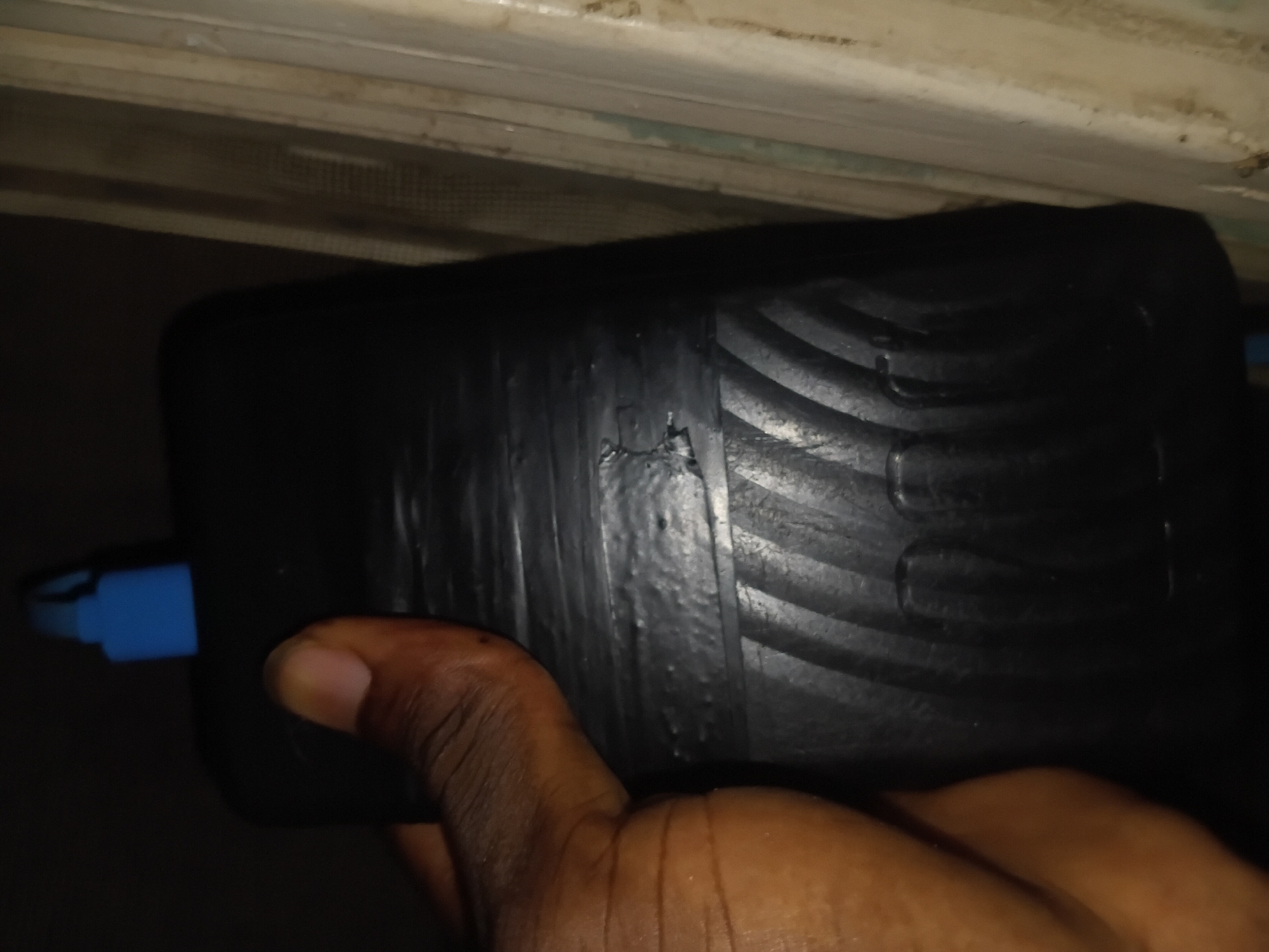 A dark-skinned hand is holding a black, cylindrical object with a ribbed texture on the bottom half. The top of the object has a blue cap. The object appears to be some kind of container. The background is dark and out of focus, with some vertical wooden elements visible on the right side. The image is undated and its location in Jalingo, Nigeria, is not discernible from the visual information provided.